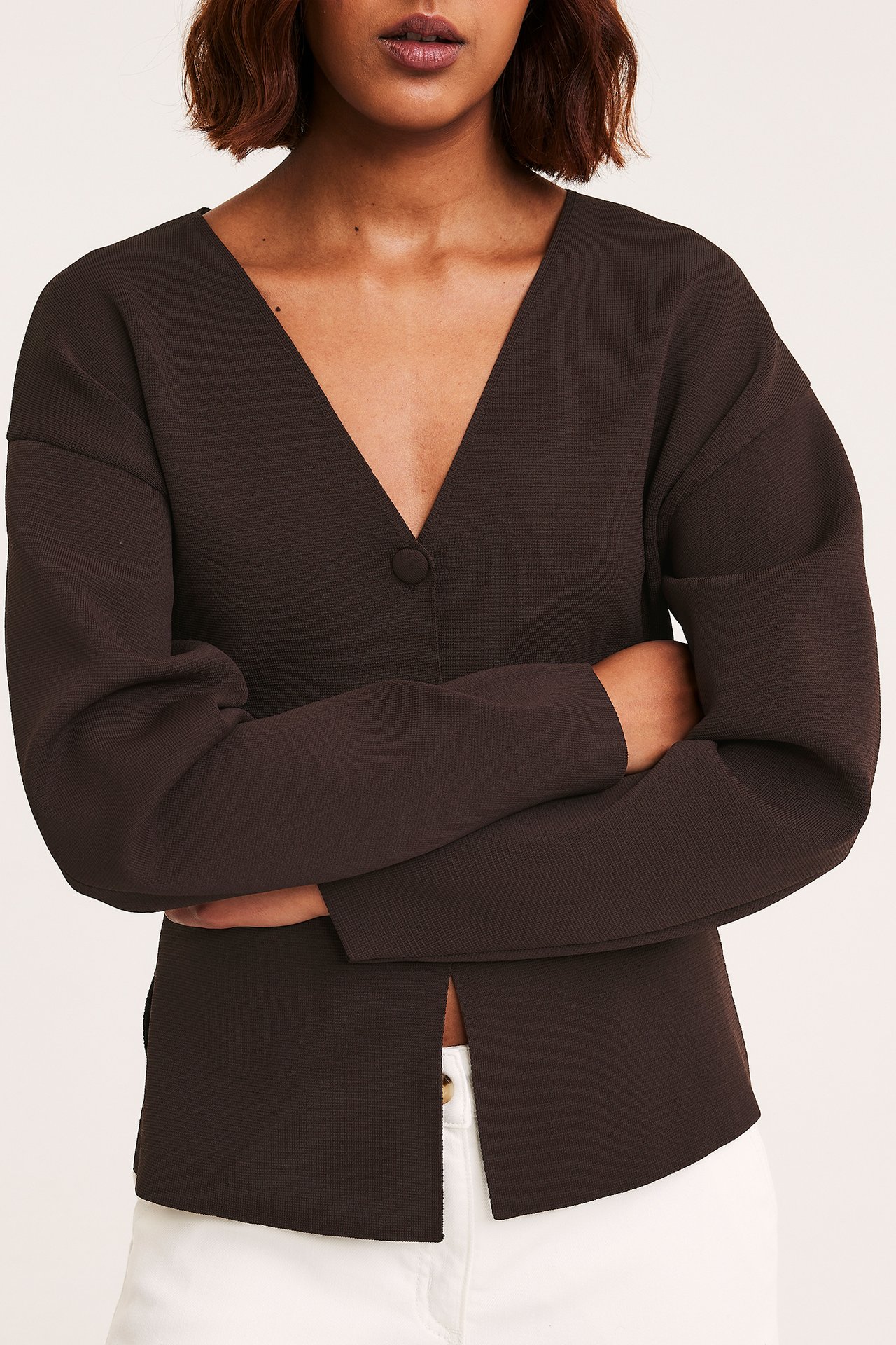 Naisen tummanruskea V-aukkoine neuletakki, yhden napin ja etuhalkion kera, valkoisten housujen kanssa.