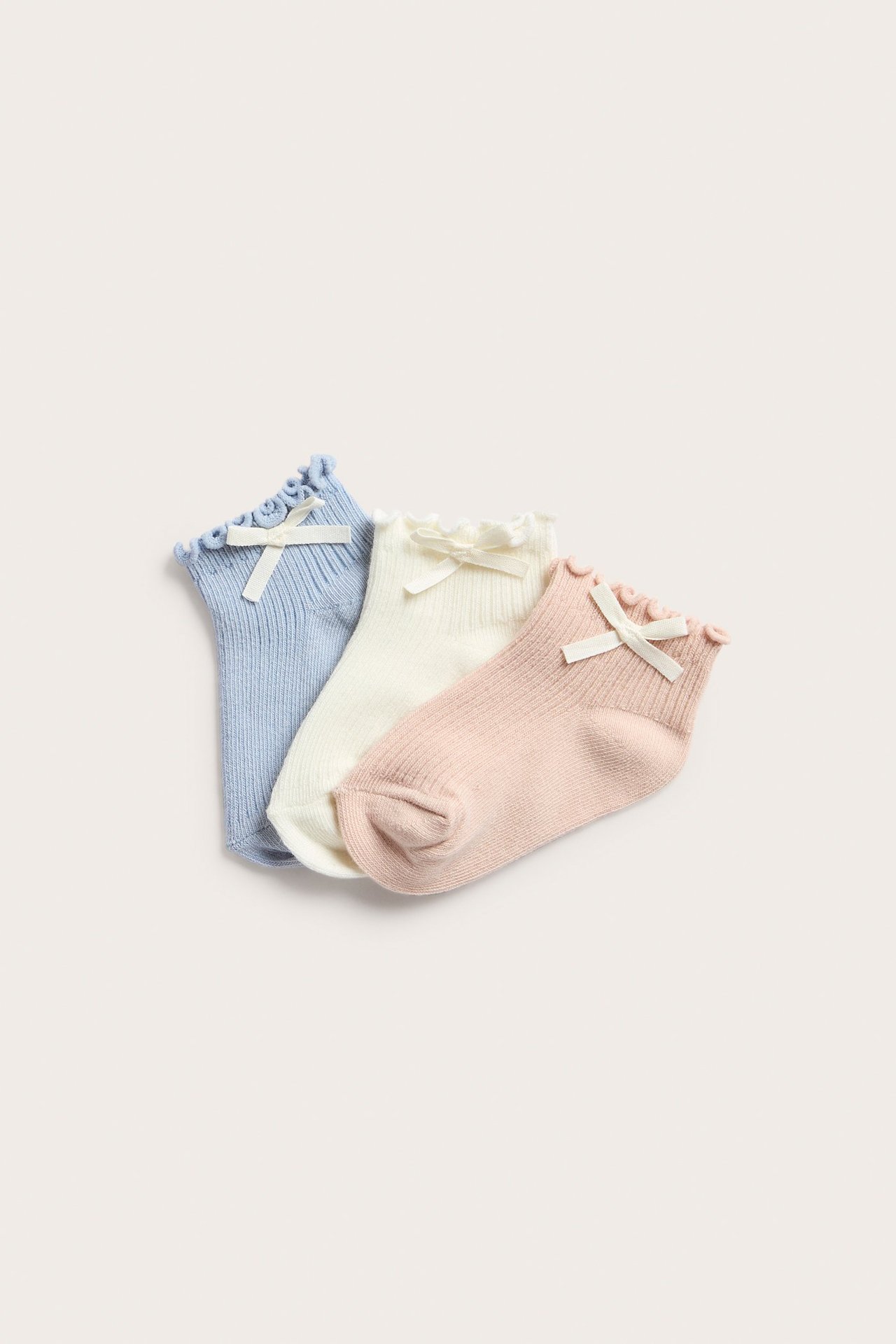 Widok z góry na trzy prążkowane krótkie skarpetki: jasnoniebieskie, ecru i jasnoróżowe, z falbankami i małymi kokardkami.