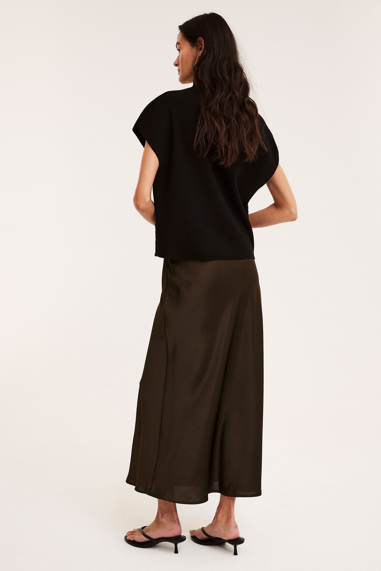 Bakfra av kvinne i mørk brun utsvunget satengskjørt, svart ermeløs topp og svarte sandaler.