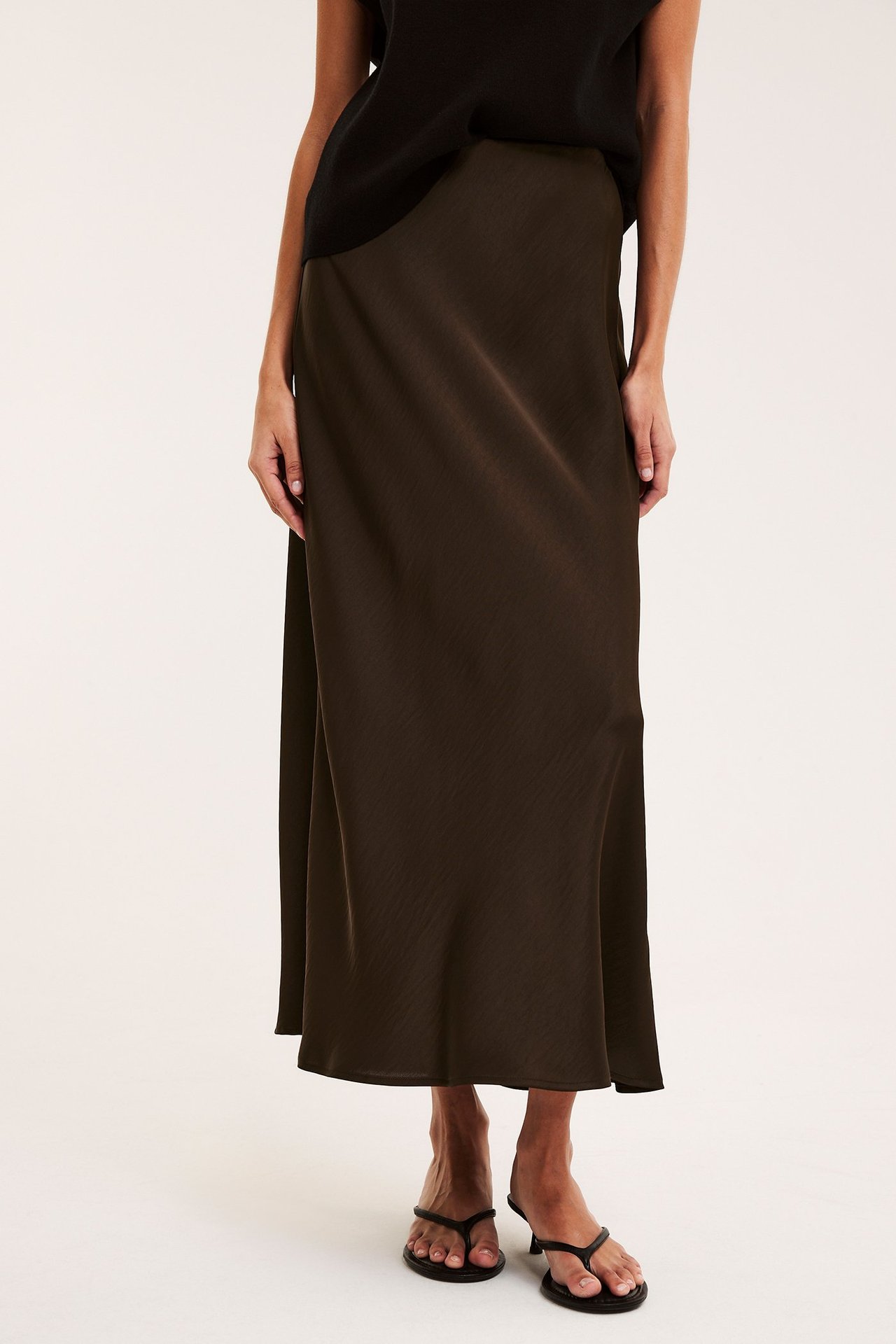 Forfra: kvinne i mørkbrun utskrådd satengskjørt, stylet med svart topp og svarte hælsandaler.