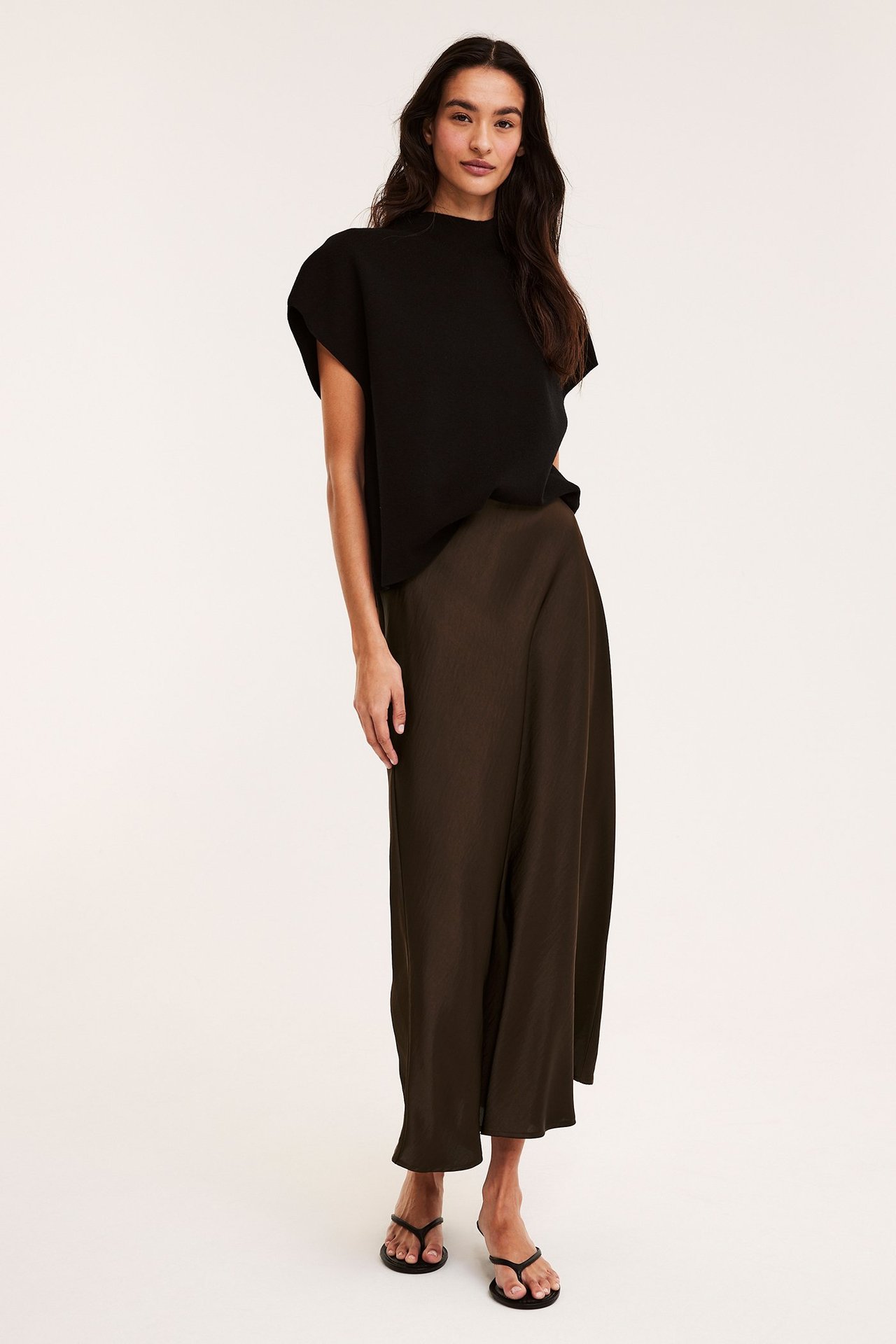 Frontbilde av kvinne i mørk brun utsvunget sateng midiskjørt, svart topp og svarte flip-flops.