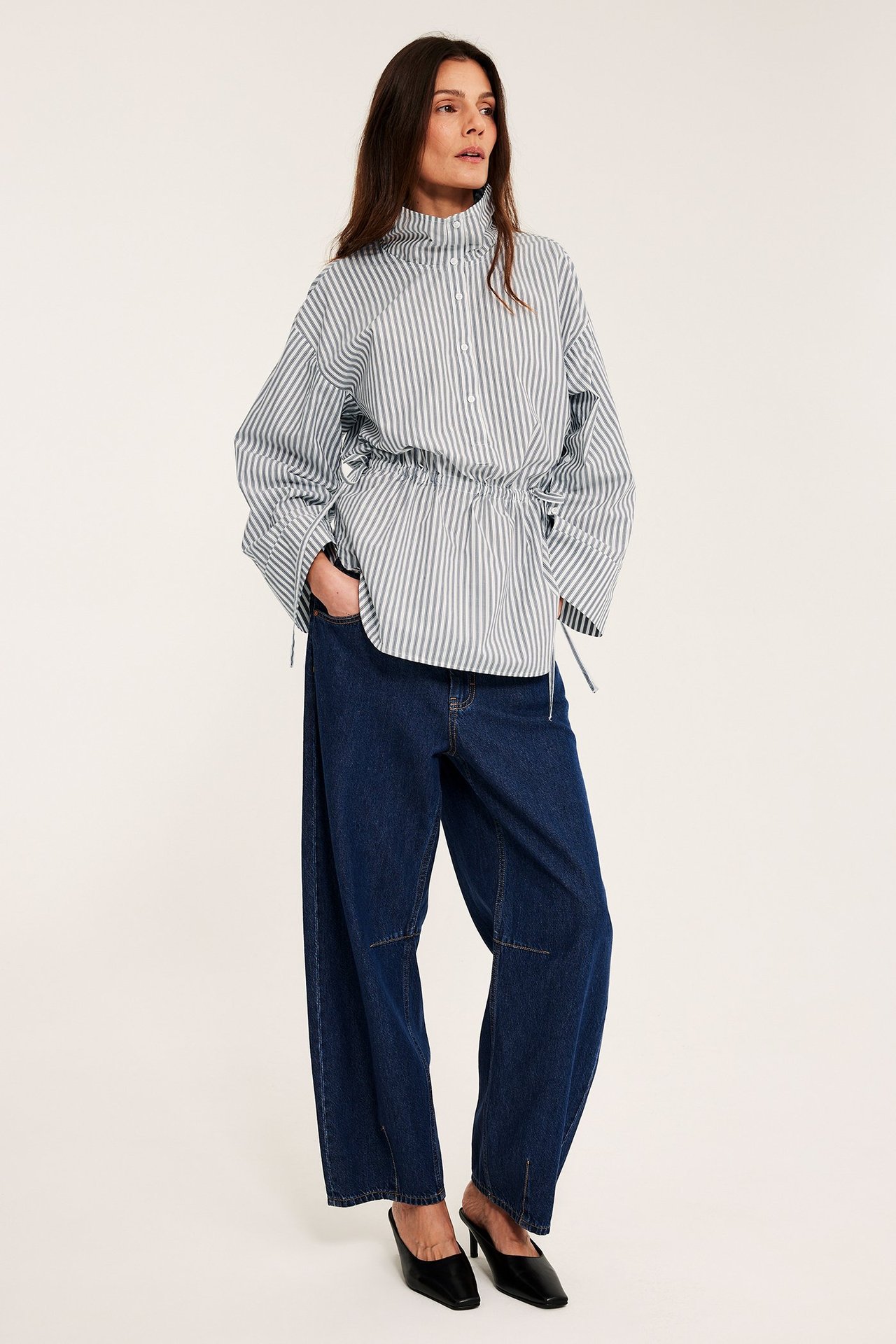 Frontvy av kvinna i offwhite- och grårandig poplinskjorta med dragsko, mörkblå vida jeans och svarta mules.