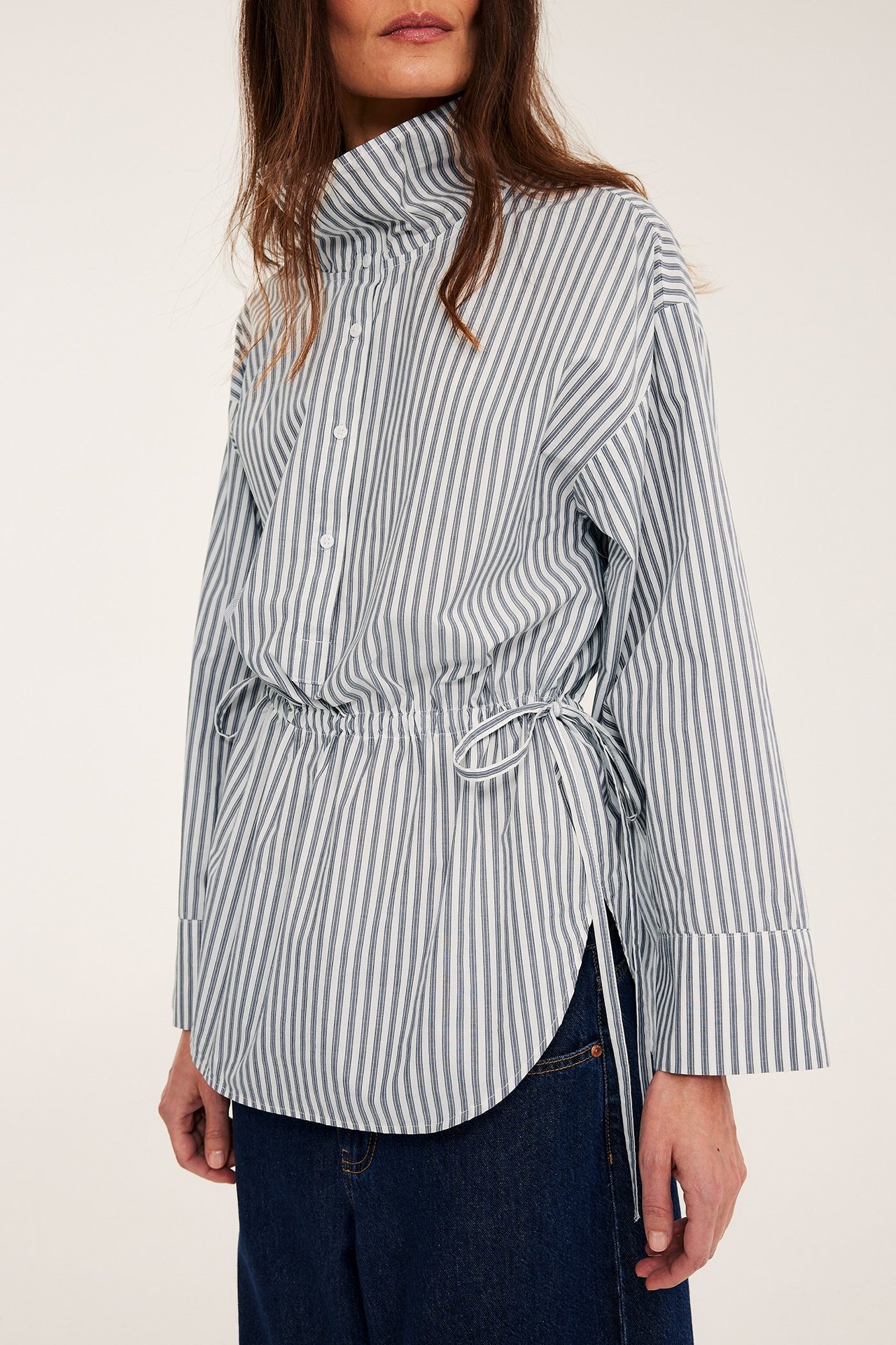 Framifrån: Kvinna bär offwhite/mörkgrå randig bomullsskjorta med dragsko i midjan, över blå jeans.