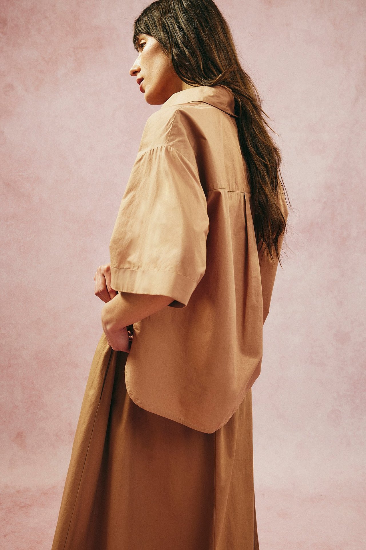 Widok z boku: Kobieta w luźnej beżowej koszuli z popeliny bawełnianej z asymetrycznym dołem i pasującą spódnicą.