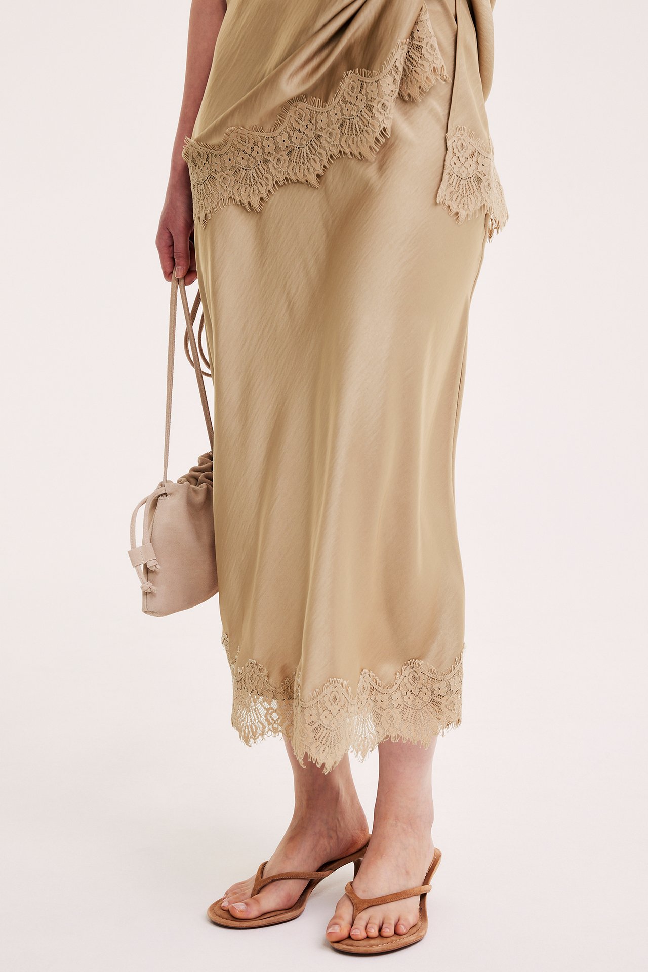 Kvinnas frontvy: Ljusbrun satinkjol med spetskant, ljusbeige dragskoväska och bruna klackade sandaler.