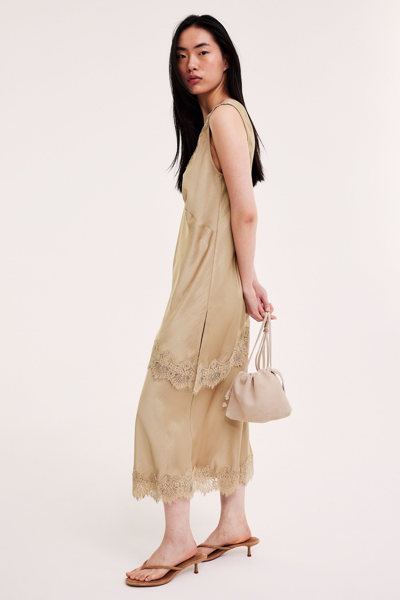 Elegancka kobieta w jasnobrązowej satynowej spódnicy z koronkowym obszyciem i gumką, trzyma beżową torebkę-worek.