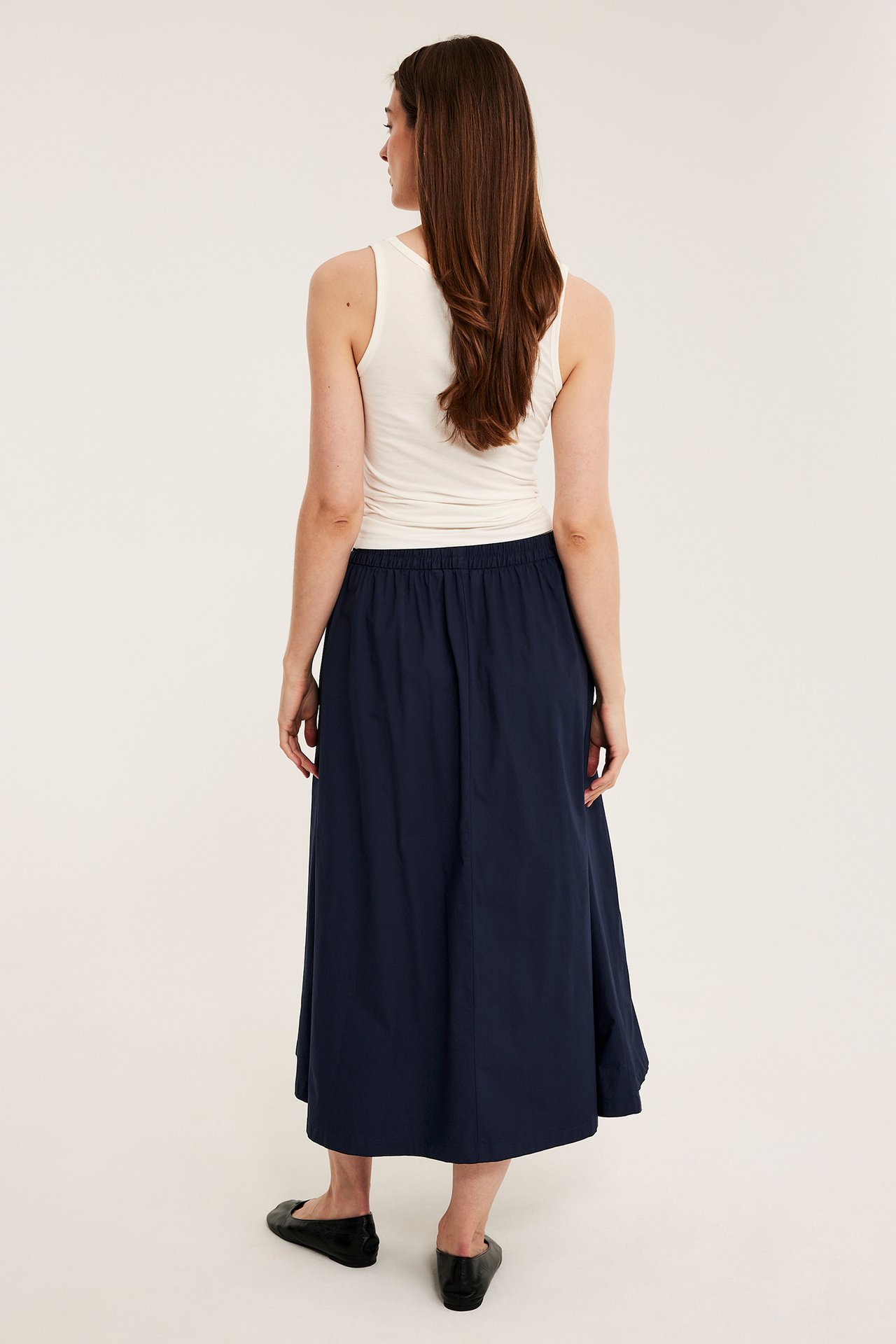 Nainen takaapäin tummansinisessä popliinihameessa, luonnonvalkoisessa topissa ja mustissa ballerinakengissä.