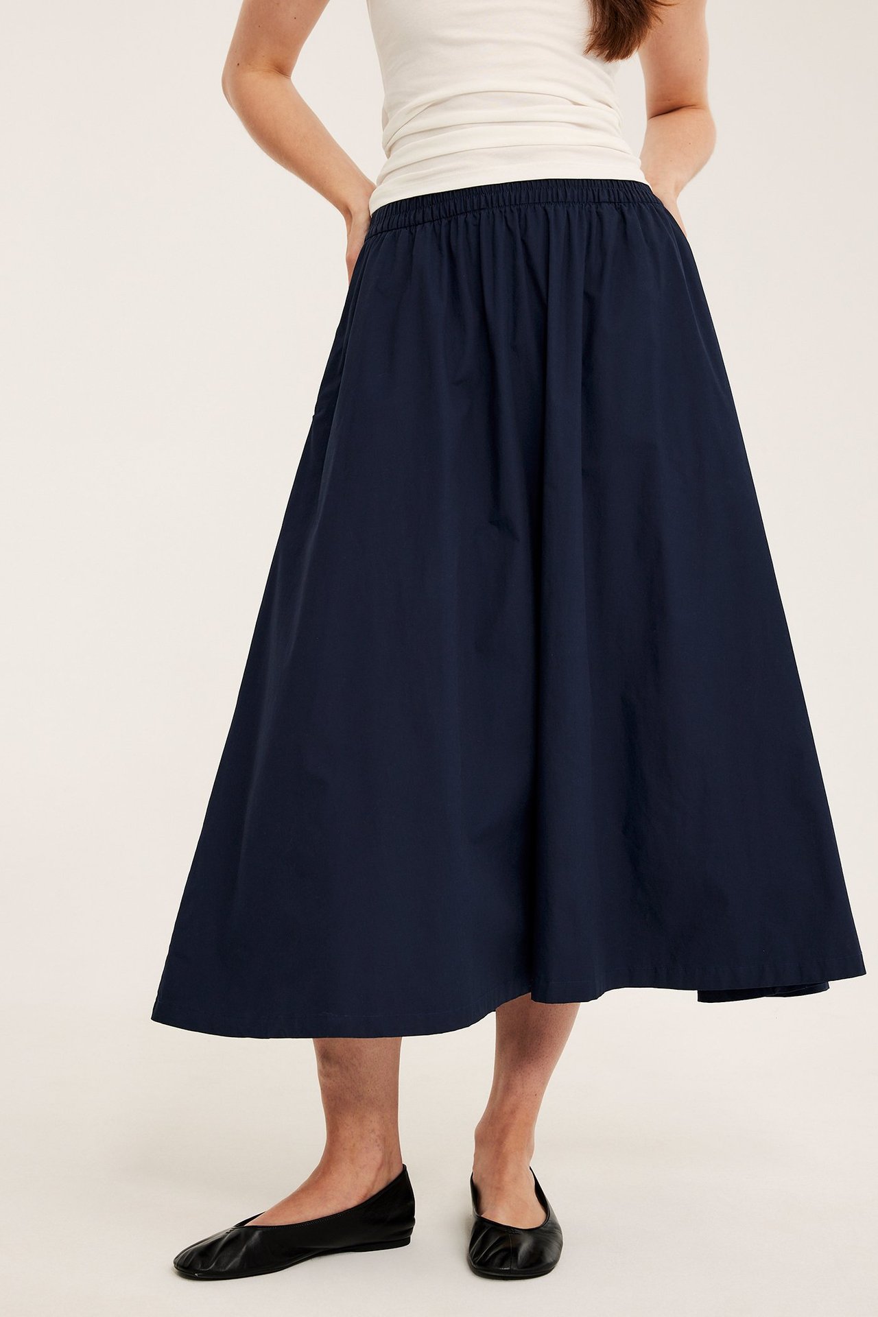 Nainen tummansinisessä levenevässä puuvillapopliinihameessa, valkoisessa topissa ja mustissa ballerinakengissä.