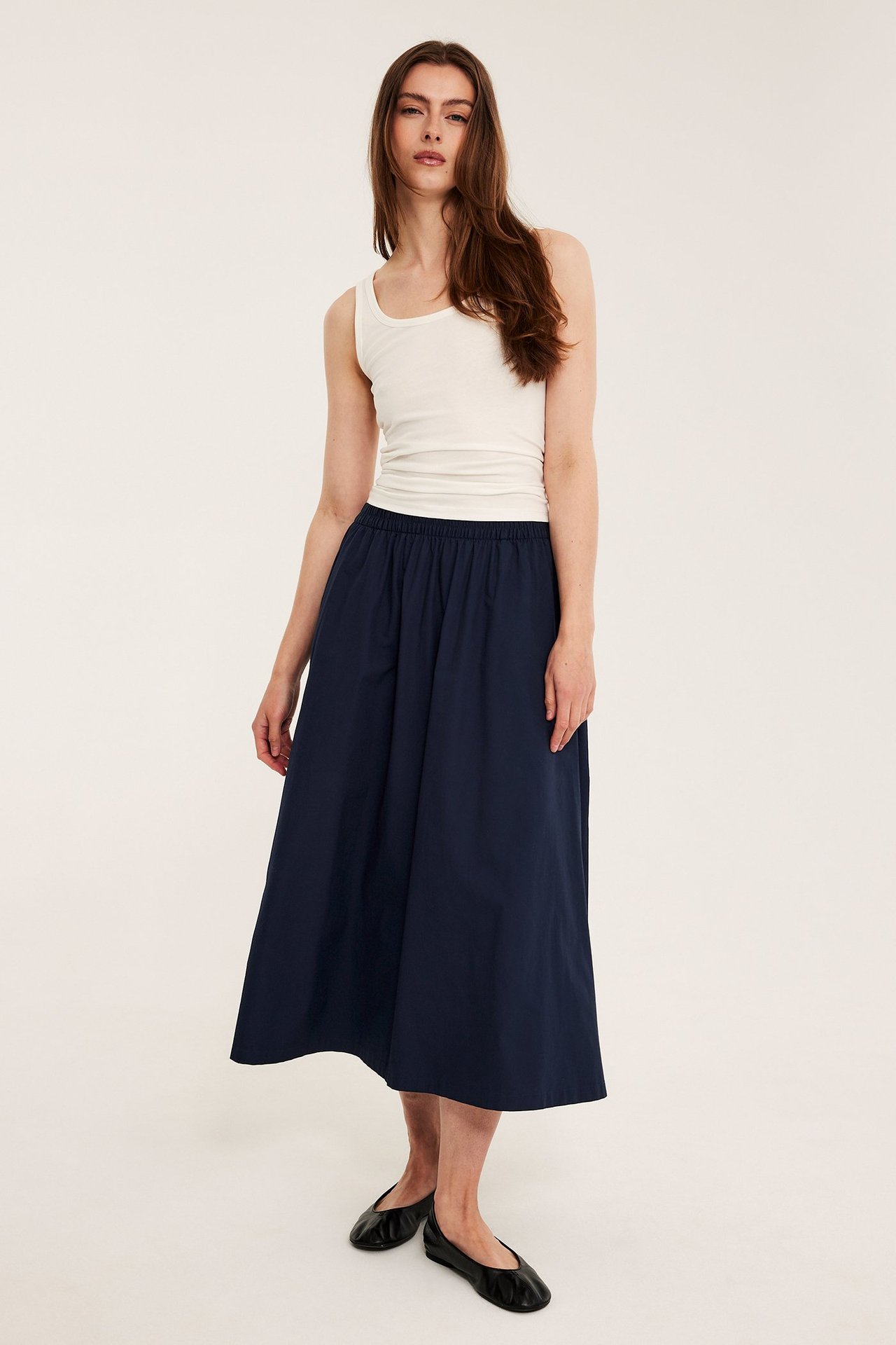 Framifrån: en kvinna i en mörkblå utställd bomullspoplinkjol, ett vitt linne och svarta platta skor.