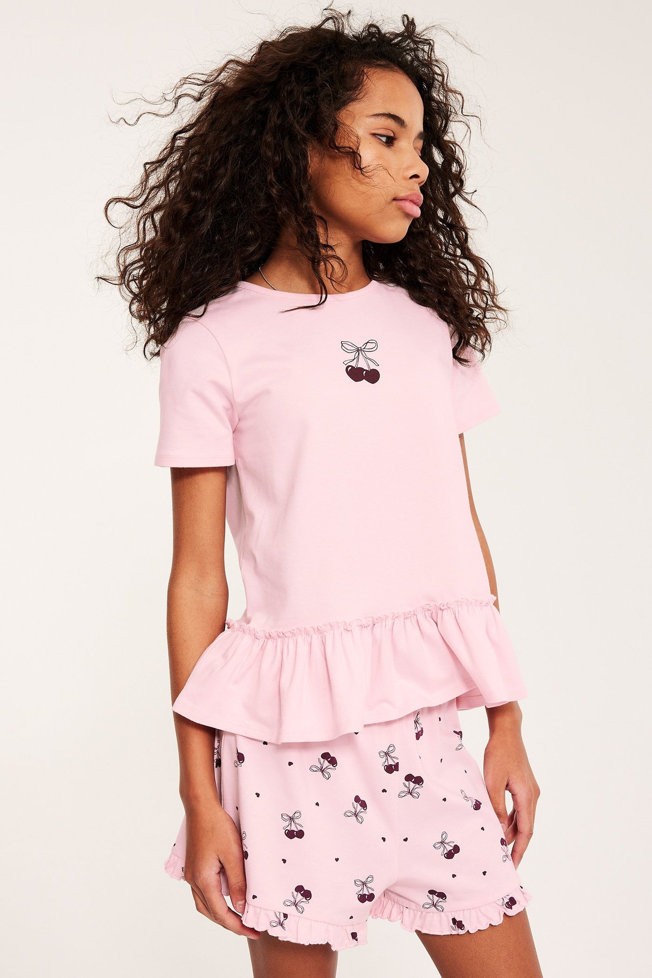 Framifrån: Barn i rosa kortärmad pyjamas med körsbärstryck och volangprydd peplumtopp.