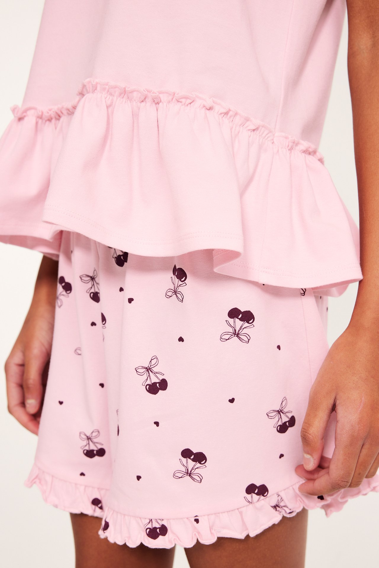 Framifrån: Kid bär rosa kortärmad pyjamas med körsbärs- och hjärtmönster, volang på topp och shorts.