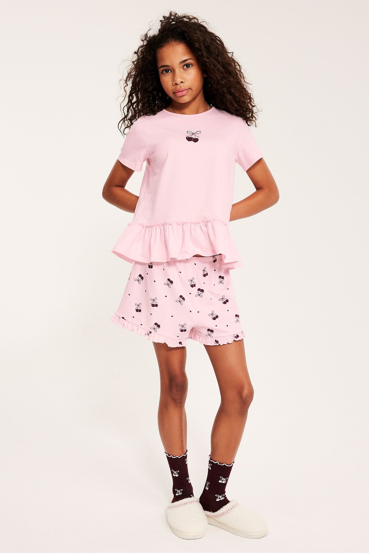 Framifrån: Kid i rosa kortärmad pyjamas med körsbärstryck, matchande shorts, mörka strumpor och tofflor.
