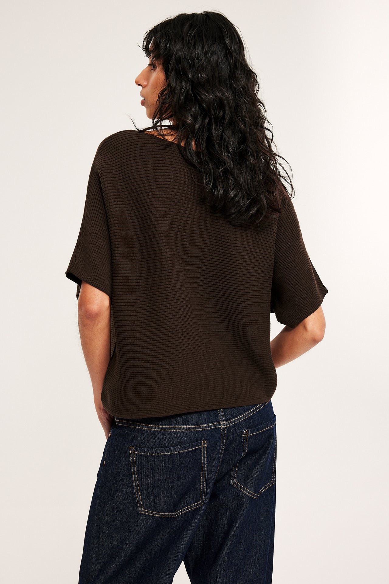 Bakfra av kvinne i mørk brun ribbestrikket kortermet genser og mørkeblå jeans.