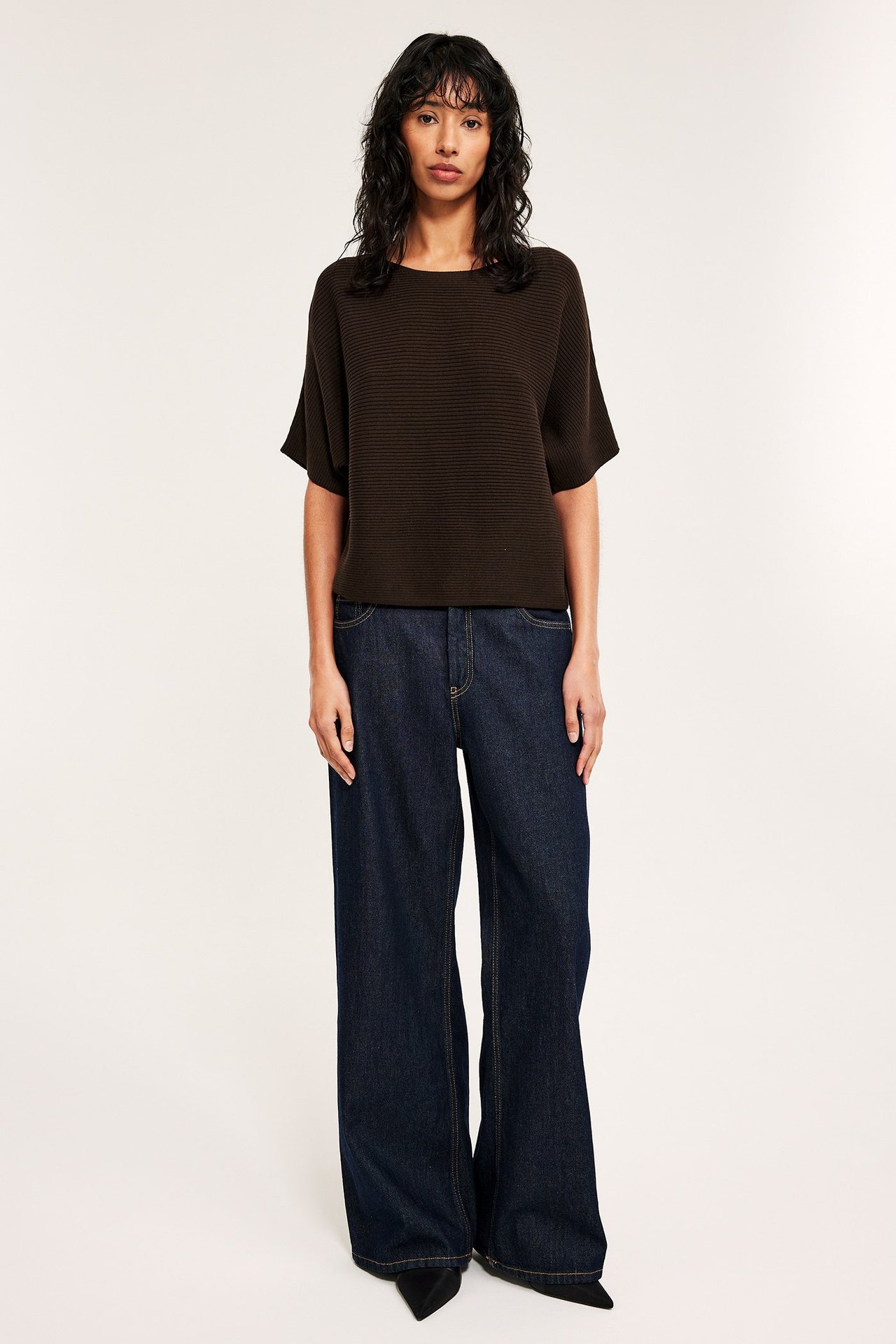 Edestä: Nainen tummanruskeassa ribatussa puuvillaneuleessa, tummansinisissä leveälahkeisissa farkuissa, mustissa kengissä.