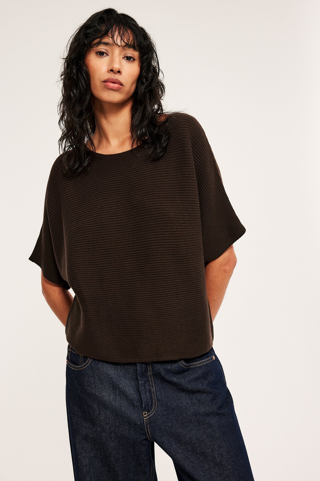 Forfra: kvinne i mørk brun ribbestrikket bomullsgenser med rund hals og mørkeblå jeans.