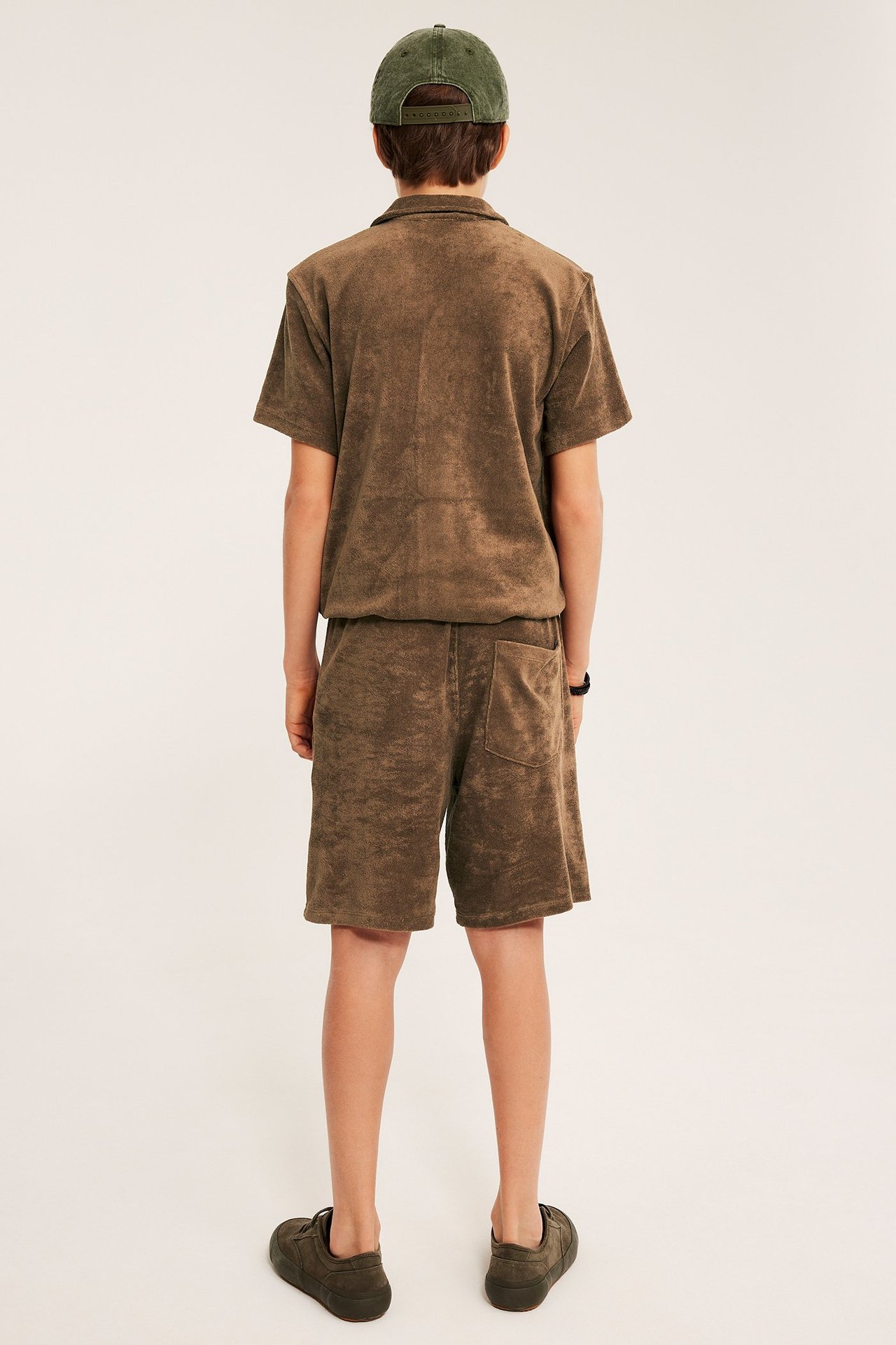 Widok z tyłu dziecka w brązowych szortach frotte, pasującej koszulce frotte, ciemnozielonej czapce i tenisówkach.