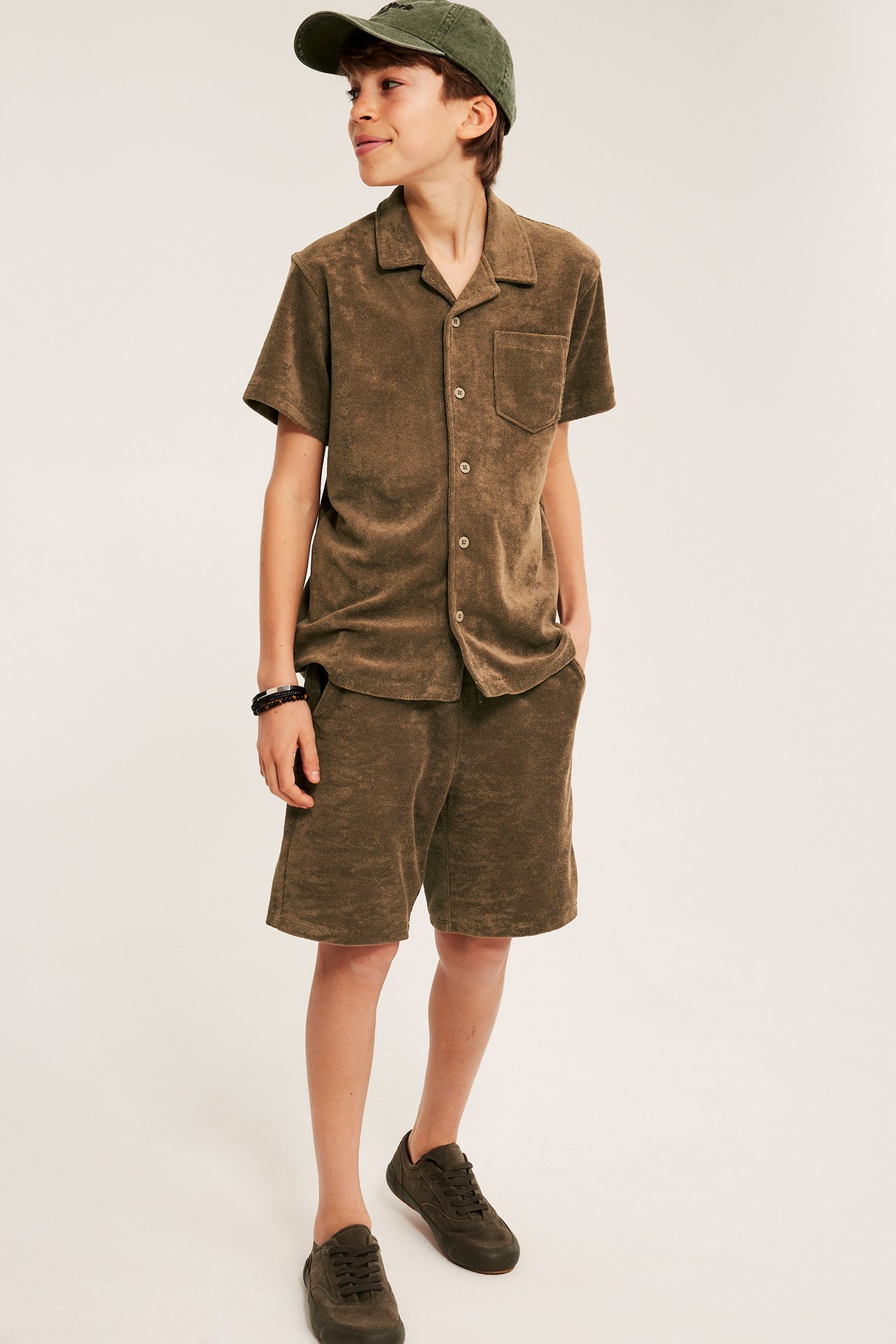 Frontbild: barn i bruna frottéshorts, matchande frottéskjorta, grön keps och bruna sneakers.