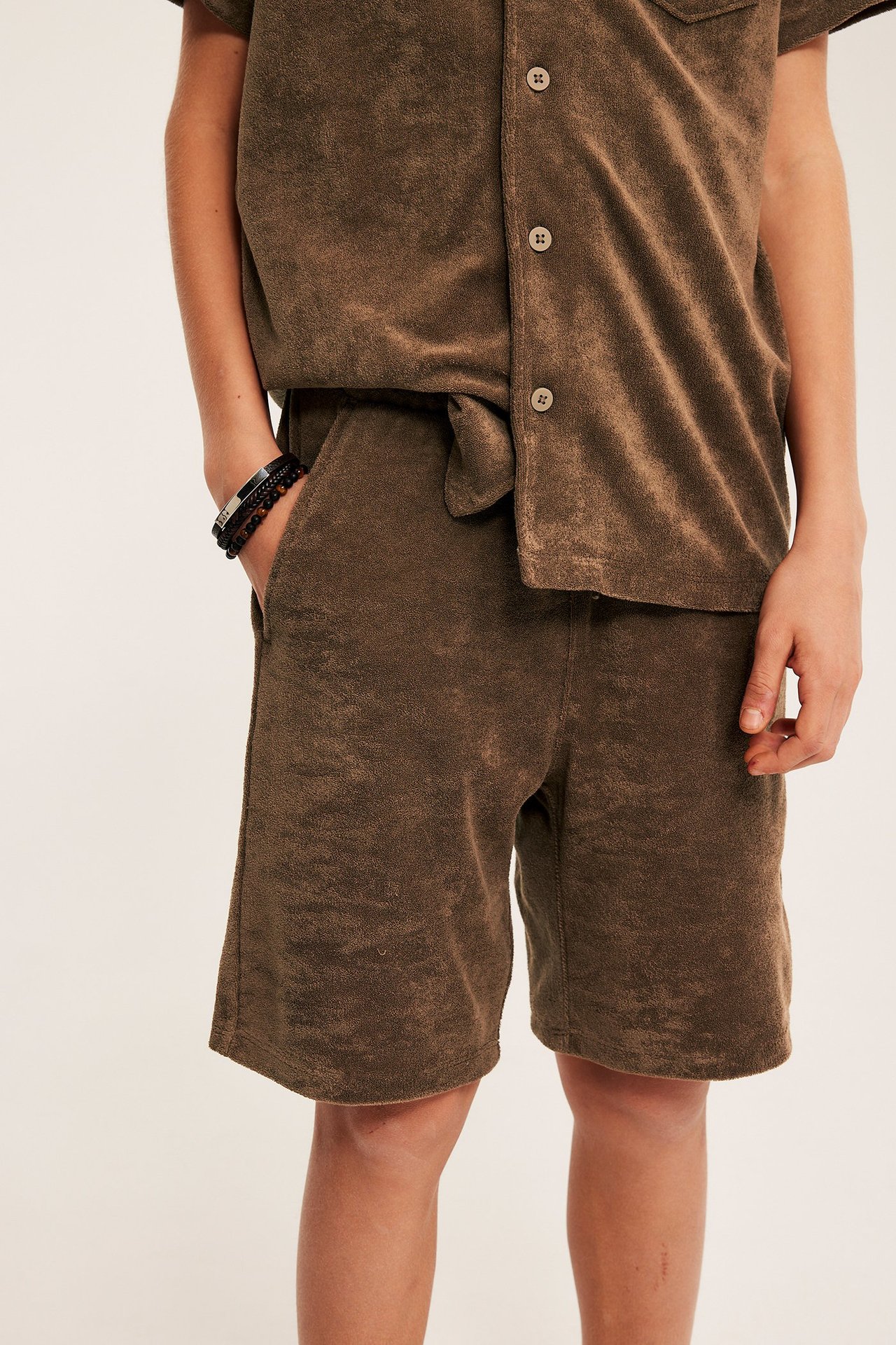 Widok z przodu dziecka w brązowych szortach frotte ze sznurkiem, dopasowanej koszulce i bransoletkach.