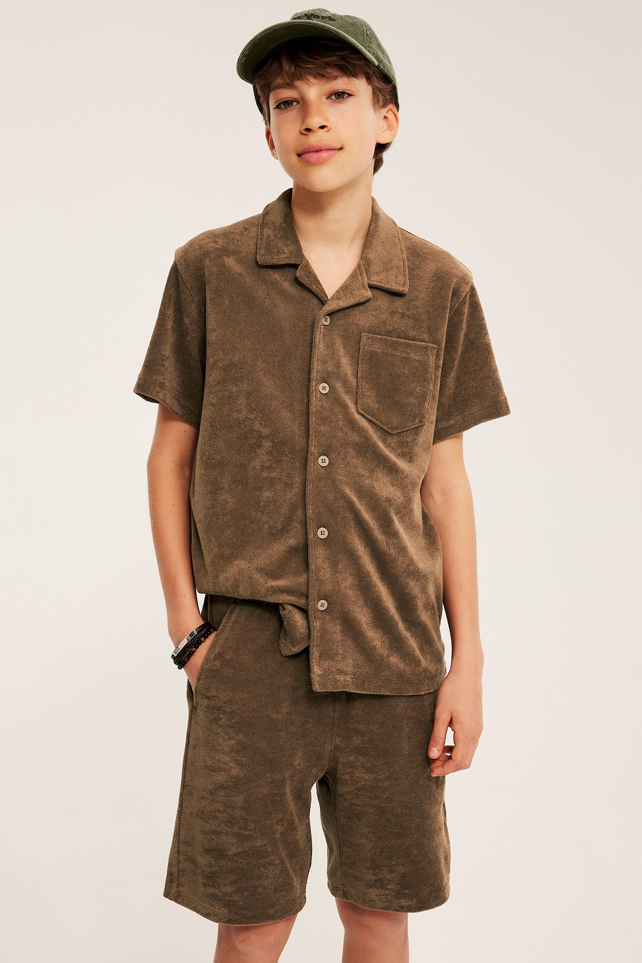 Frontvy av en pojke i bruna frottéshorts med dragsko, matchande skjorta och en grön keps.