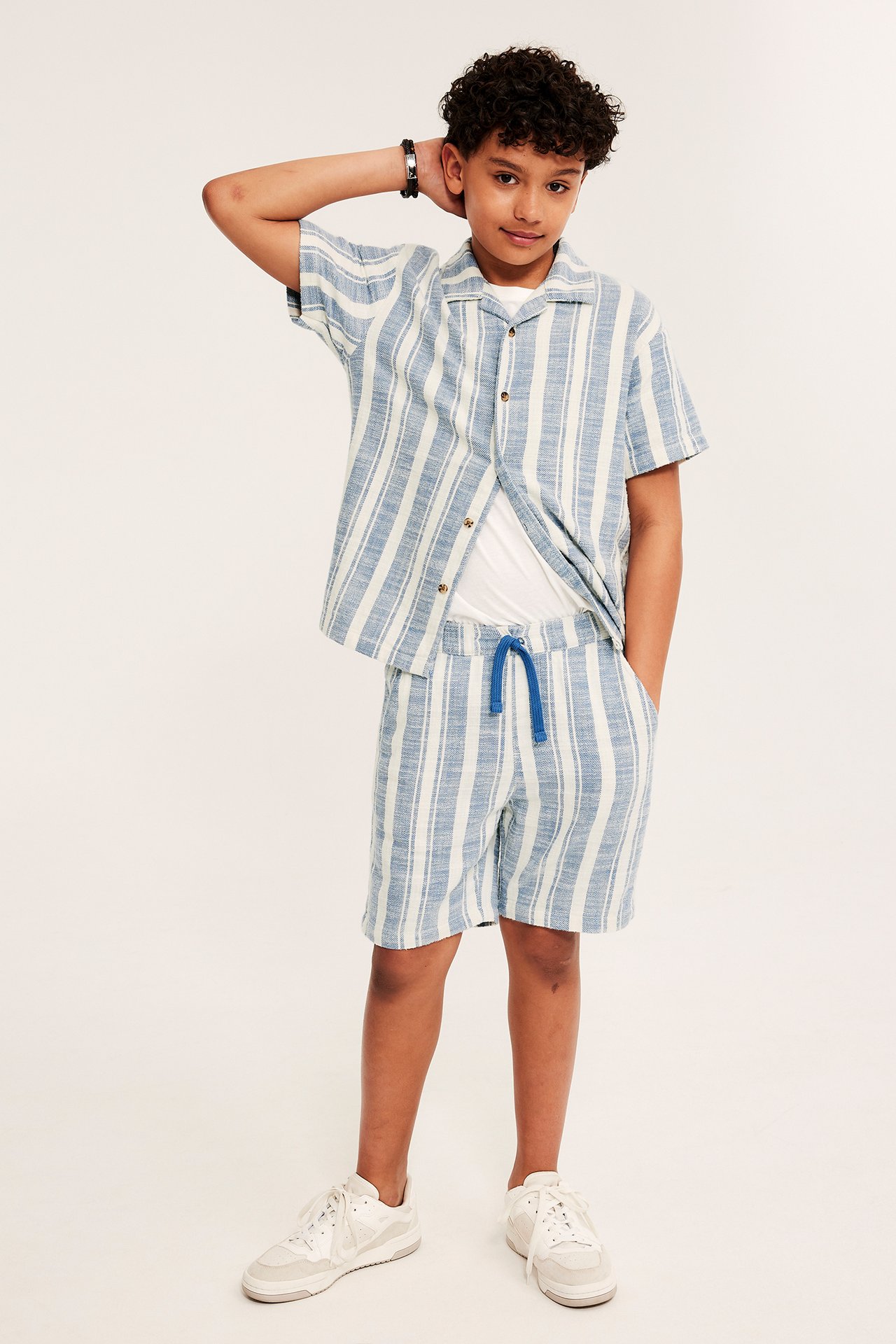 Widok z przodu: Dziecko w niebieskich szortach w paski ze sznurkiem, otwartej koszuli i białym t-shircie.
