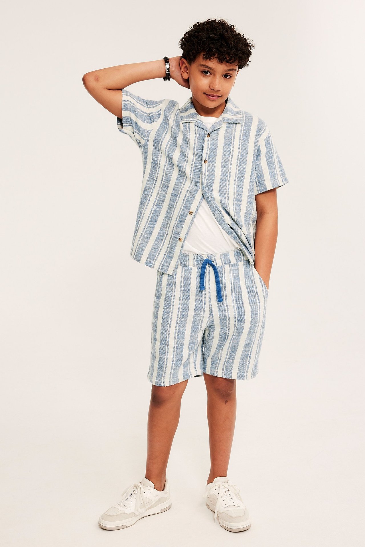 Widok z przodu: Dziecko w niebieskich szortach w paski ze sznurkiem, otwartej koszuli i białym t-shircie.
