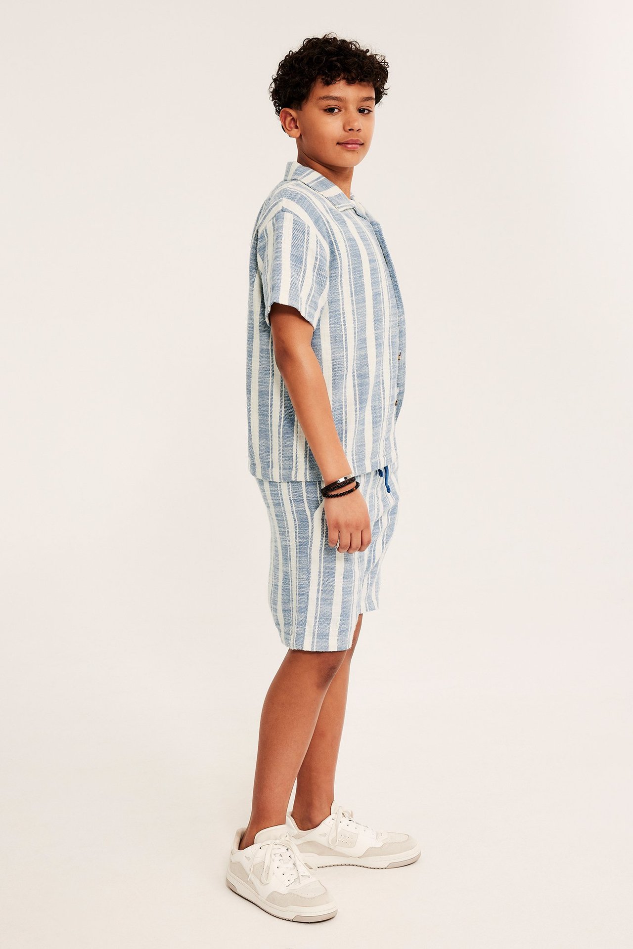 Widok z boku: Dziecko w niebiesko-białych bawełnianych szortach w paski, pasującej koszuli i białych sneakersach.