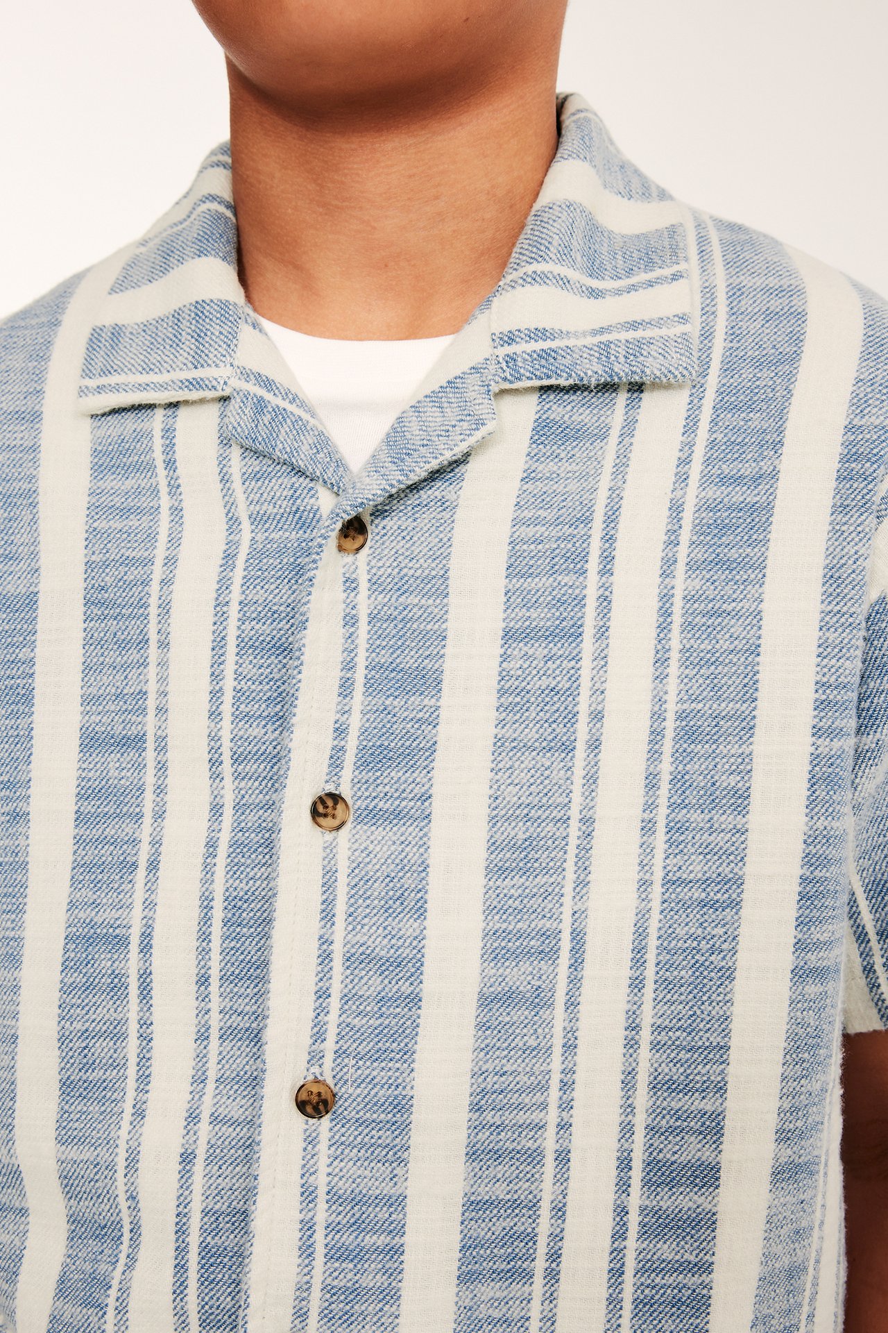 Zbliżenie z przodu dziecka w niebiesko-białej koszuli bawełnianej w paski z kołnierzem resortowym, na białej koszulce.