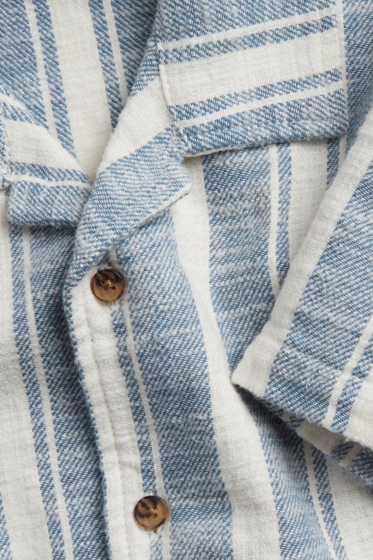 Zbliżenie: niebiesko-biała bawełniana koszula w paski, z resortowym kołnierzykiem i brązowymi guzikami.