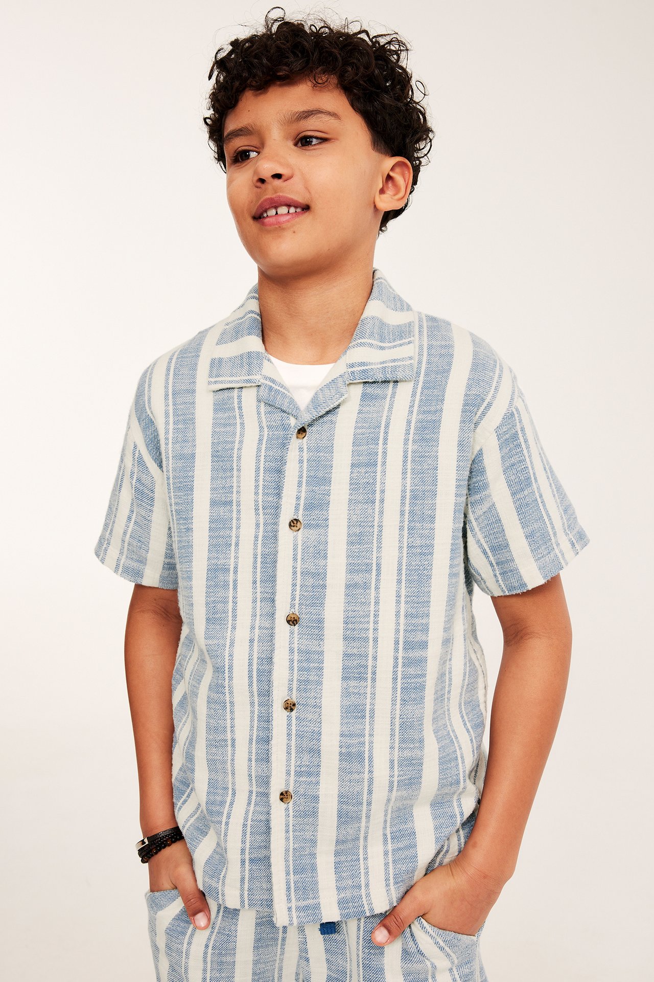 Widok z przodu: Dziecko w niebiesko-białej bawełnianej koszuli w paski, białym T-shircie, szortach, czarnej bransoletce.