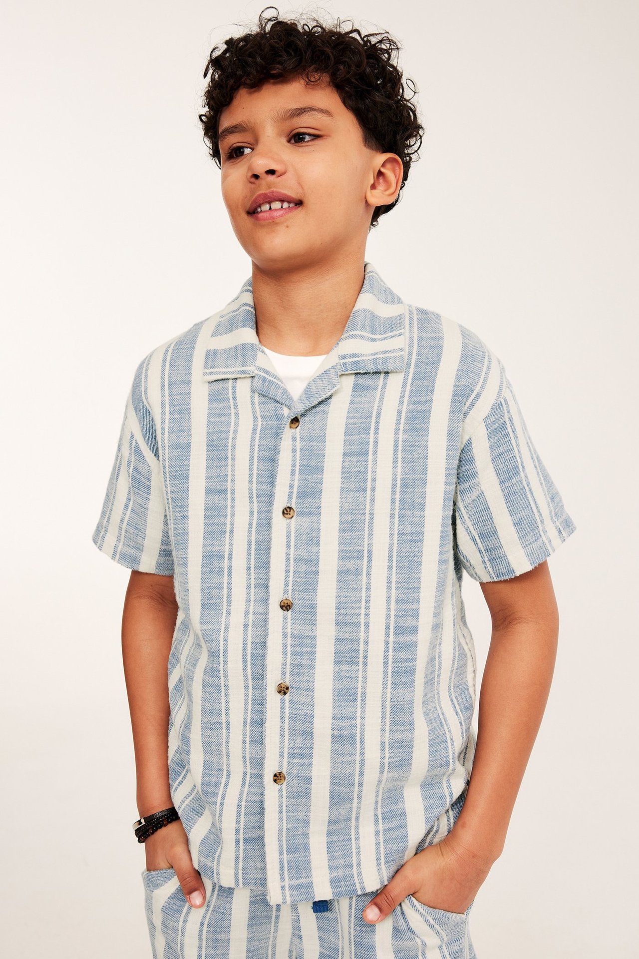 Widok z przodu: Dziecko w niebiesko-białej bawełnianej koszuli w paski, białym T-shircie, szortach, czarnej bransoletce.