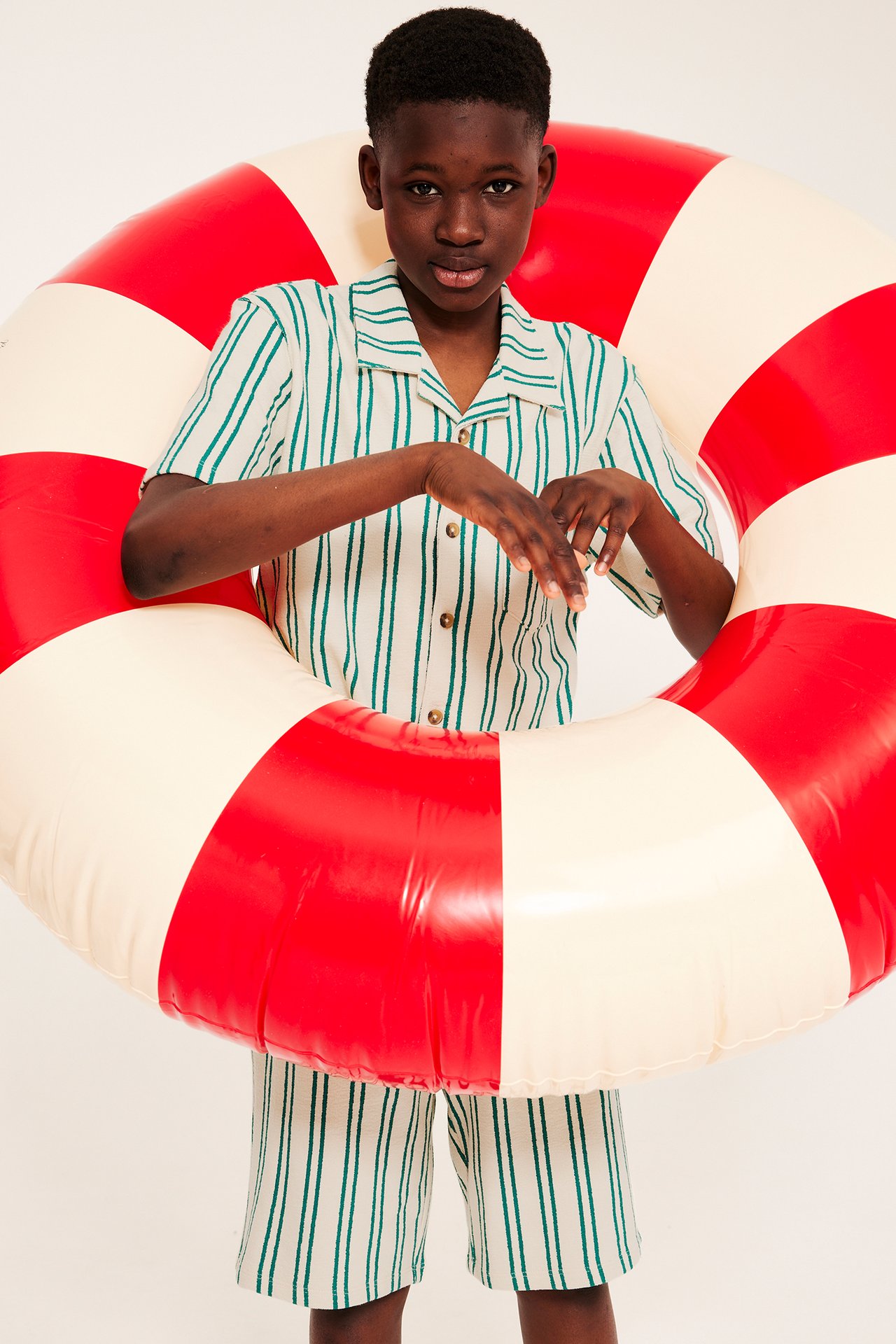 Widok z przodu: Dziecko w biało-zielonej koszuli i szortach w paski z bawełny, trzymające koło ratunkowe.