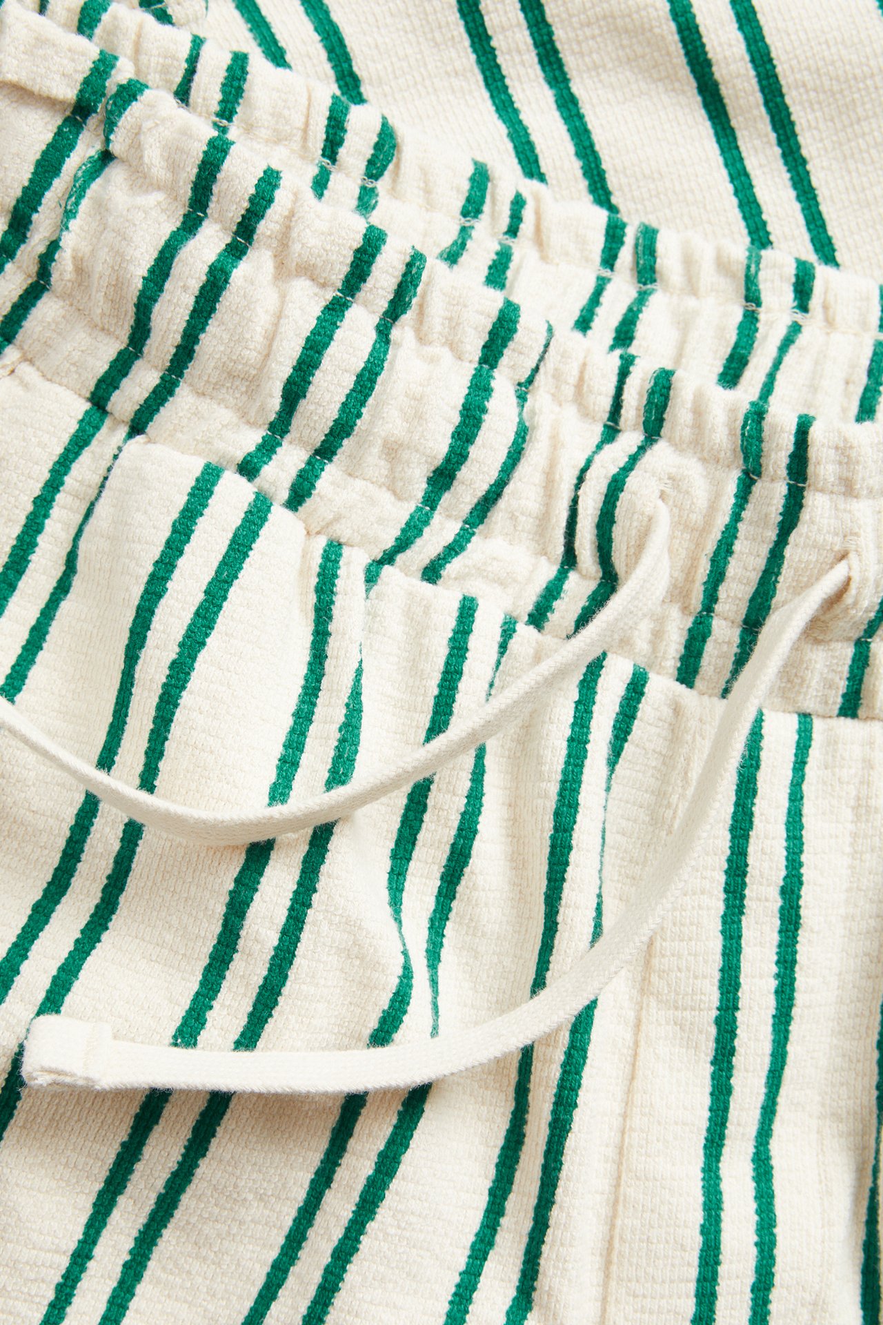 Närbild av cremefärgade bomullsstickade shorts med gröna ränder, med resårmidja och dragsko.