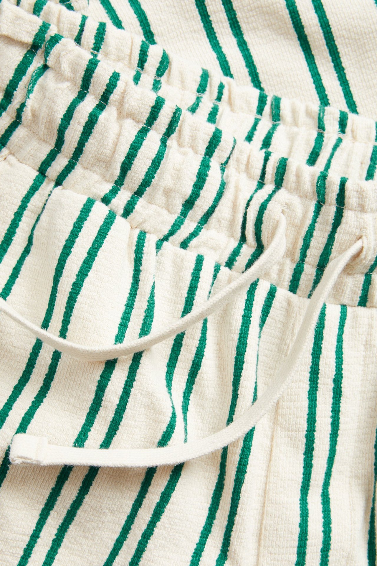 Lähikuva kermanvärisistä puuvillaneuloshortseista vihreillä raidoilla, joustavalla vyötäröllä ja kiristysnyörillä.