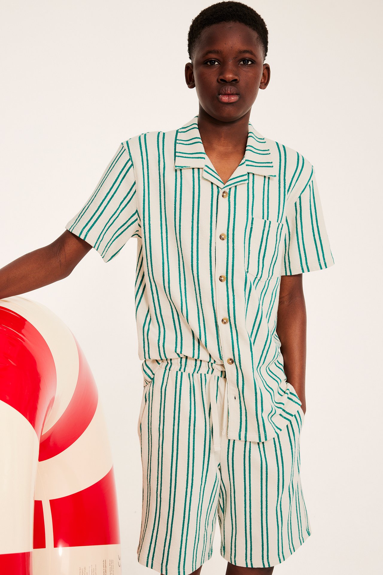 Frontvy av ett barn i gräddvit och grönrandig kortärmad skjorta samt matchande shorts i bomullstrikå.