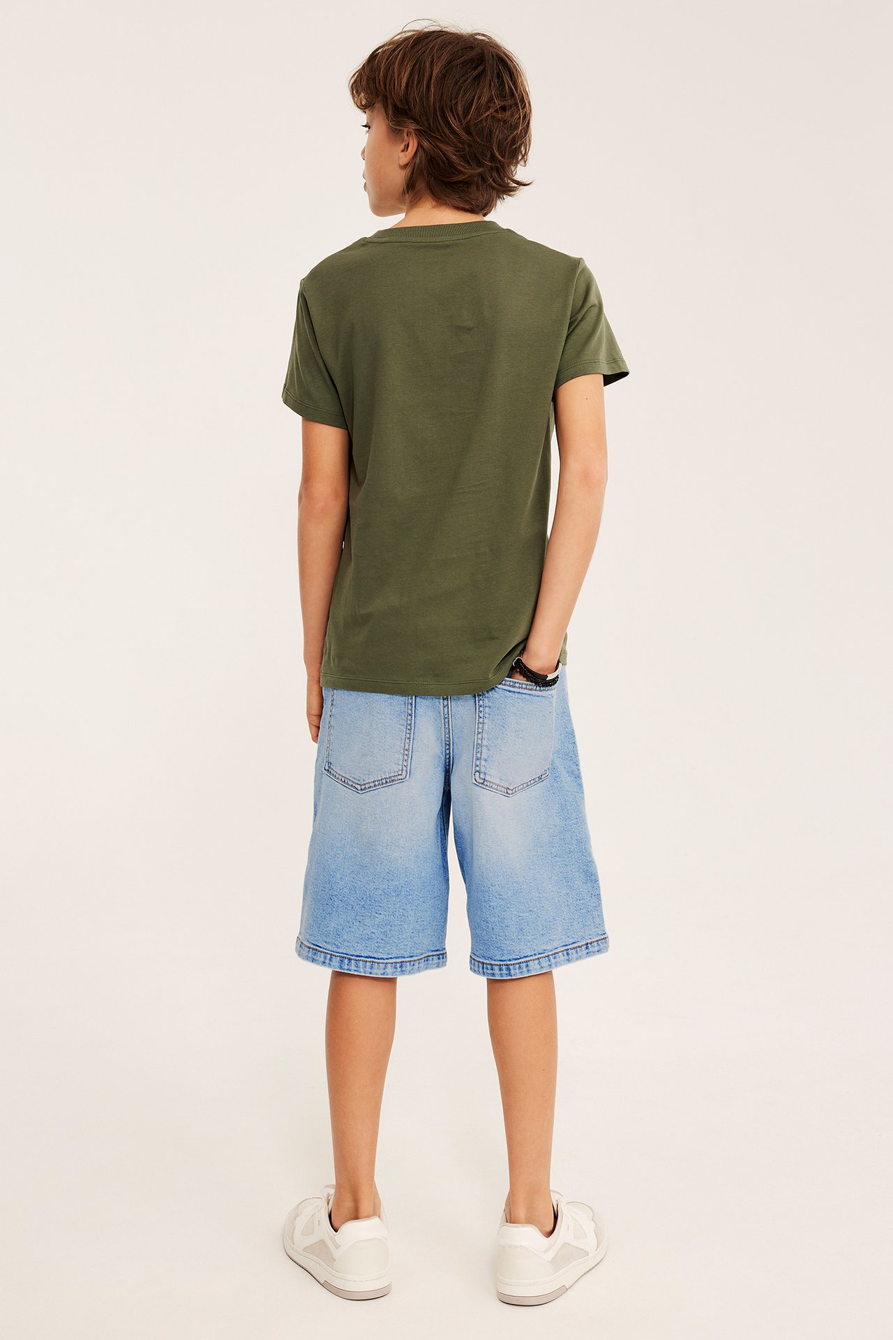 Widok z tyłu dziecka w jasnych dżinsowych szortach bermudach, oliwkowej koszulce i białych trampkach.