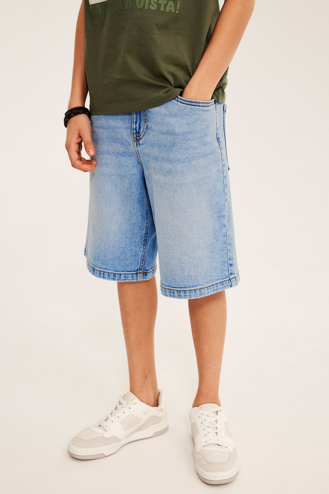 Framifrån: Barn bär ljusblå stretch denim bermudashorts, grön t-shirt, vita sneakers, svart armband.
