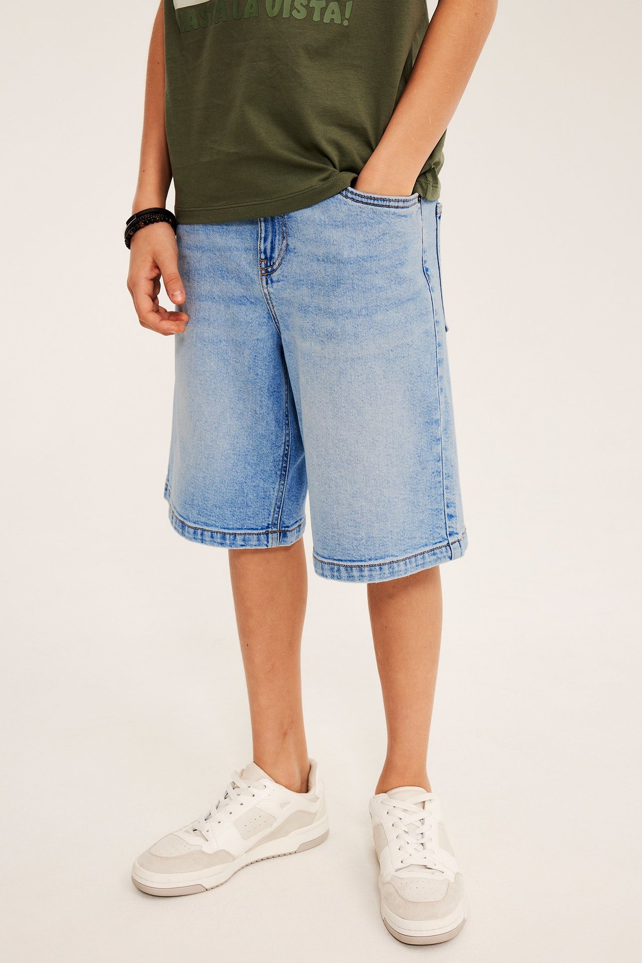 Front: Barn bruker lyseblå stretch denim bermudashorts, grønn t-skjorte, hvite joggesko, svart armbånd.