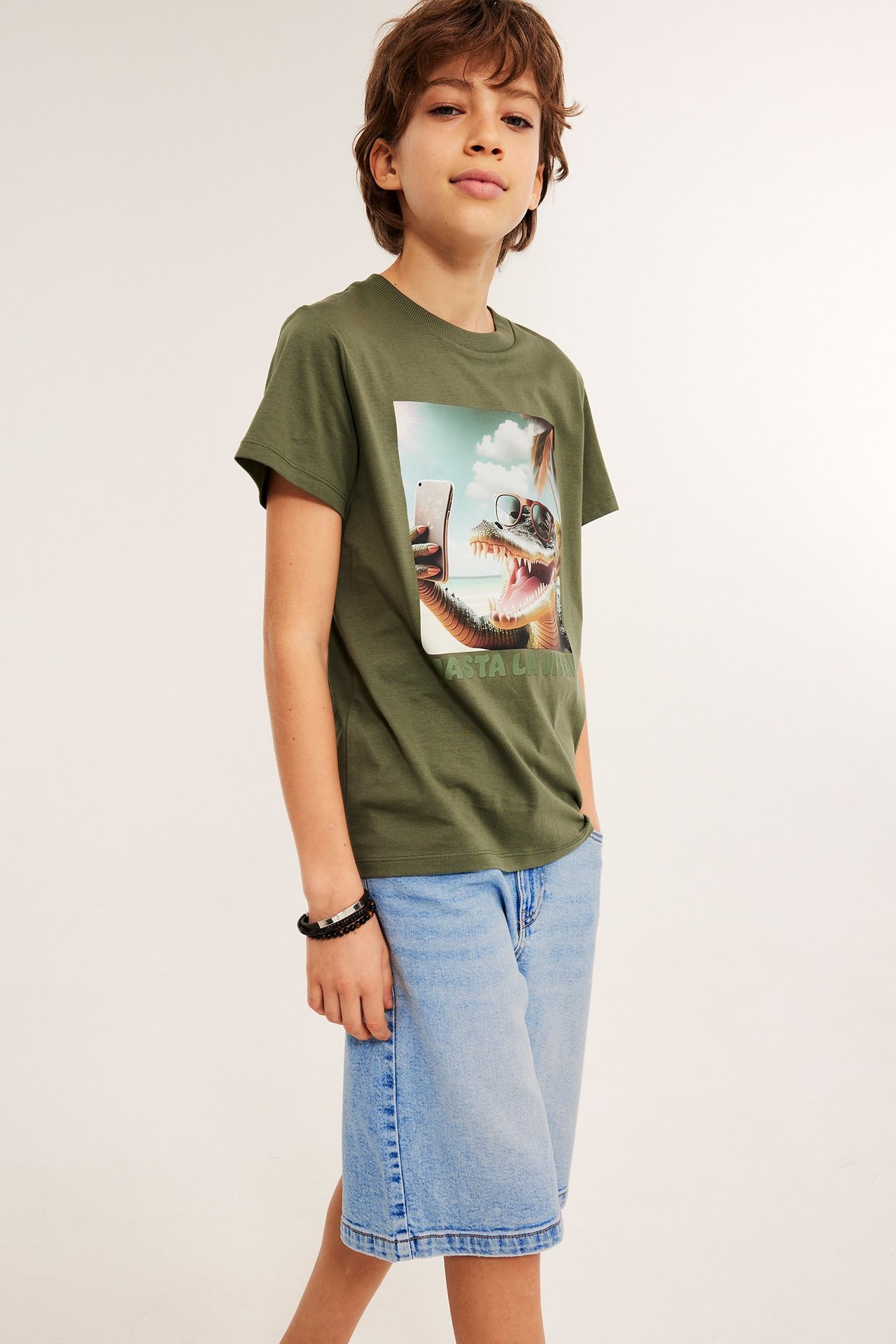 Widok z przodu: Dziecko w jasnych szortach jeansowych bermuda z guzikiem, oliwkowej koszulce z aligatorem i czarnej bransoletce.