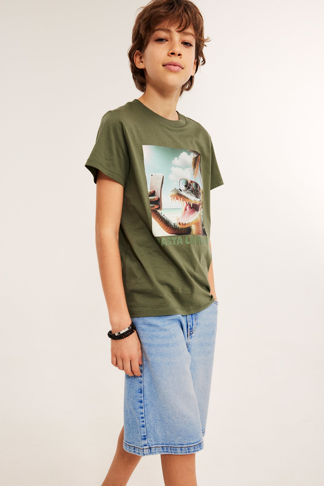 Frontvy: Barn i ljusa jeansshorts med knapp, olivgrön t-shirt med alligatortryck och ett svart armband.