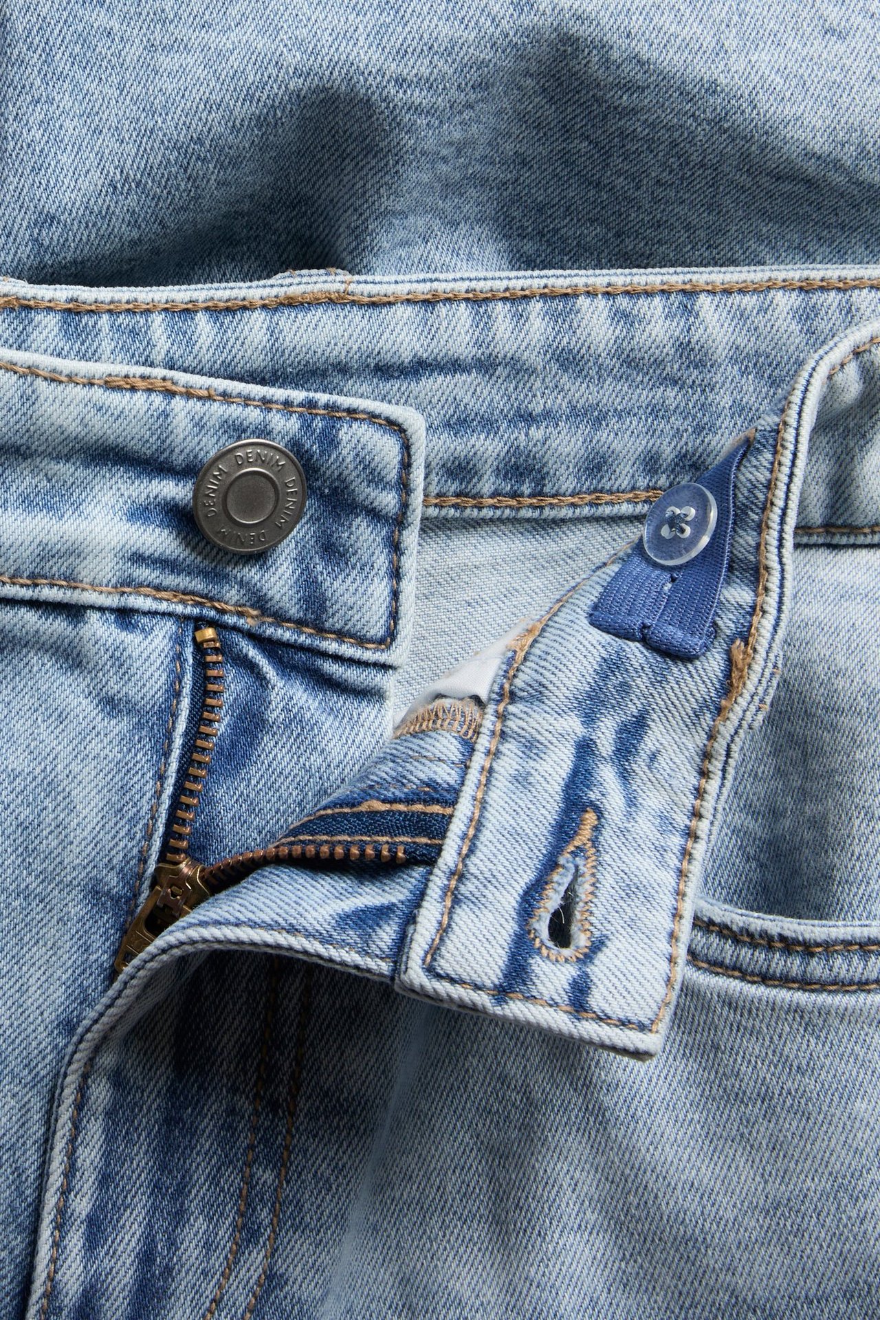 Nærbilde av lyseblå denimshorts med metallknapp, glidelås og innvendig justerbar elastisk linning.