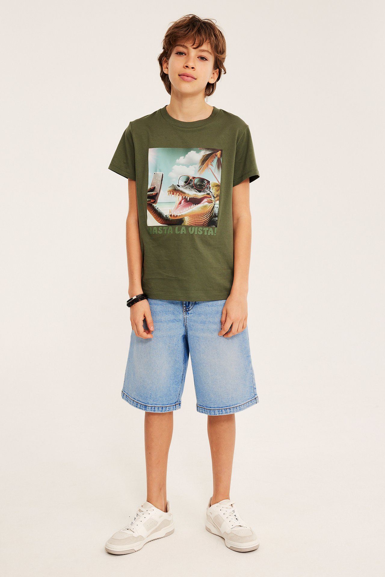Framifrån: Kid bär ljusblå denim bermudashorts, khakigrön alligator T-shirt och vita sneakers.