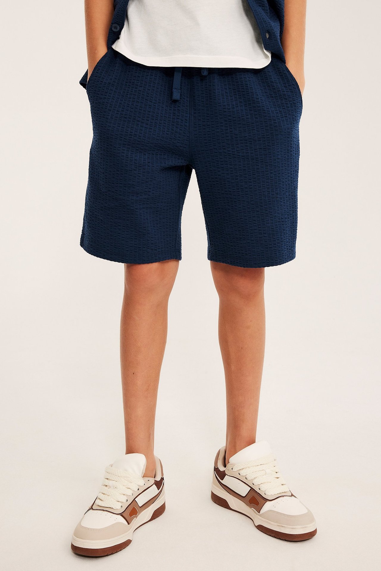 Dziecięce granatowe szorty seersucker z troczkami, biały t-shirt, granatowy kardigan i beżowe trampki.