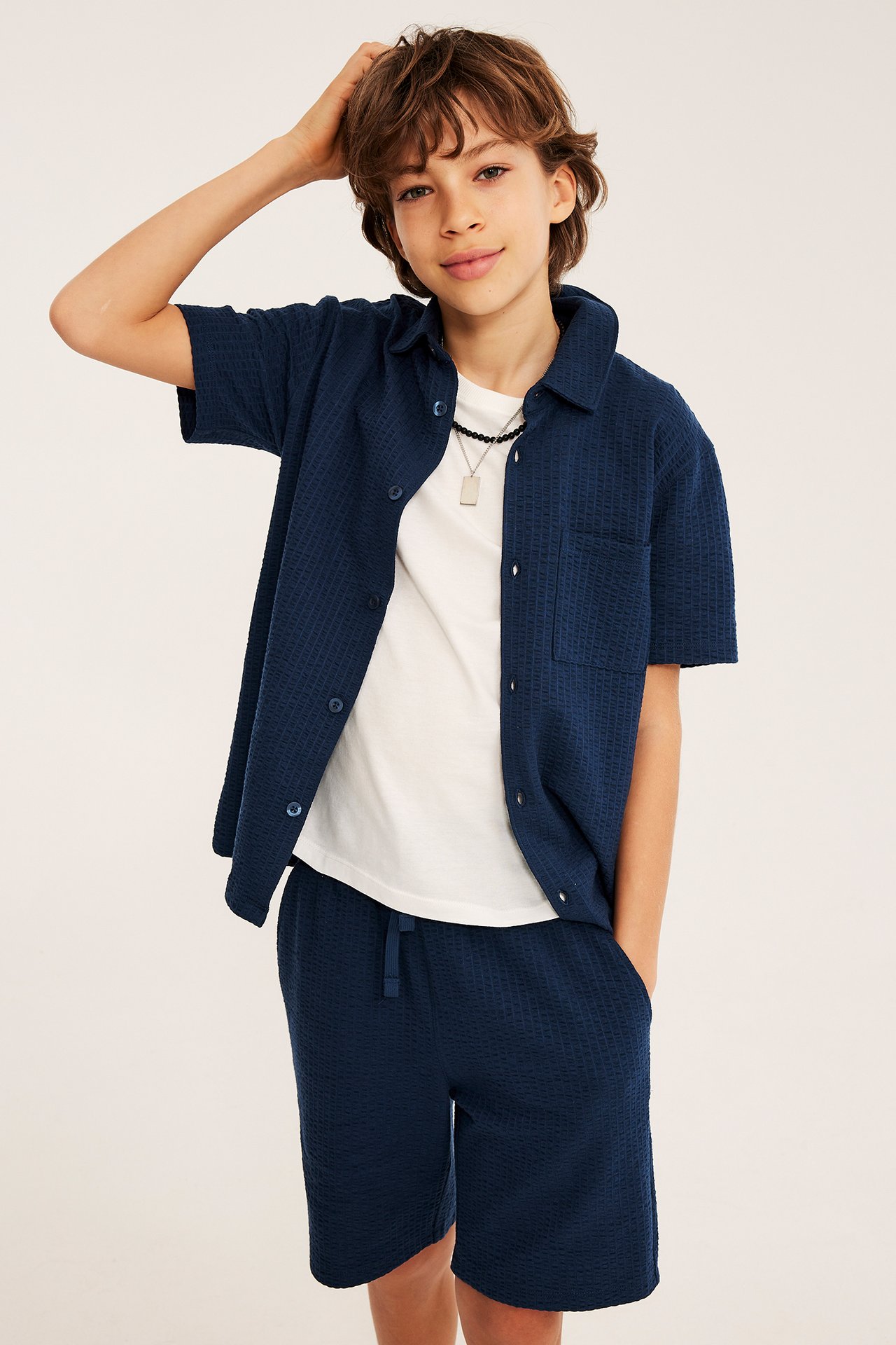 Edestä: Lapsi tummansinisessä seersucker-paidassa, yhteensopivissa shortsissa, valkoisessa t-paidassa ja kaulakorussa.