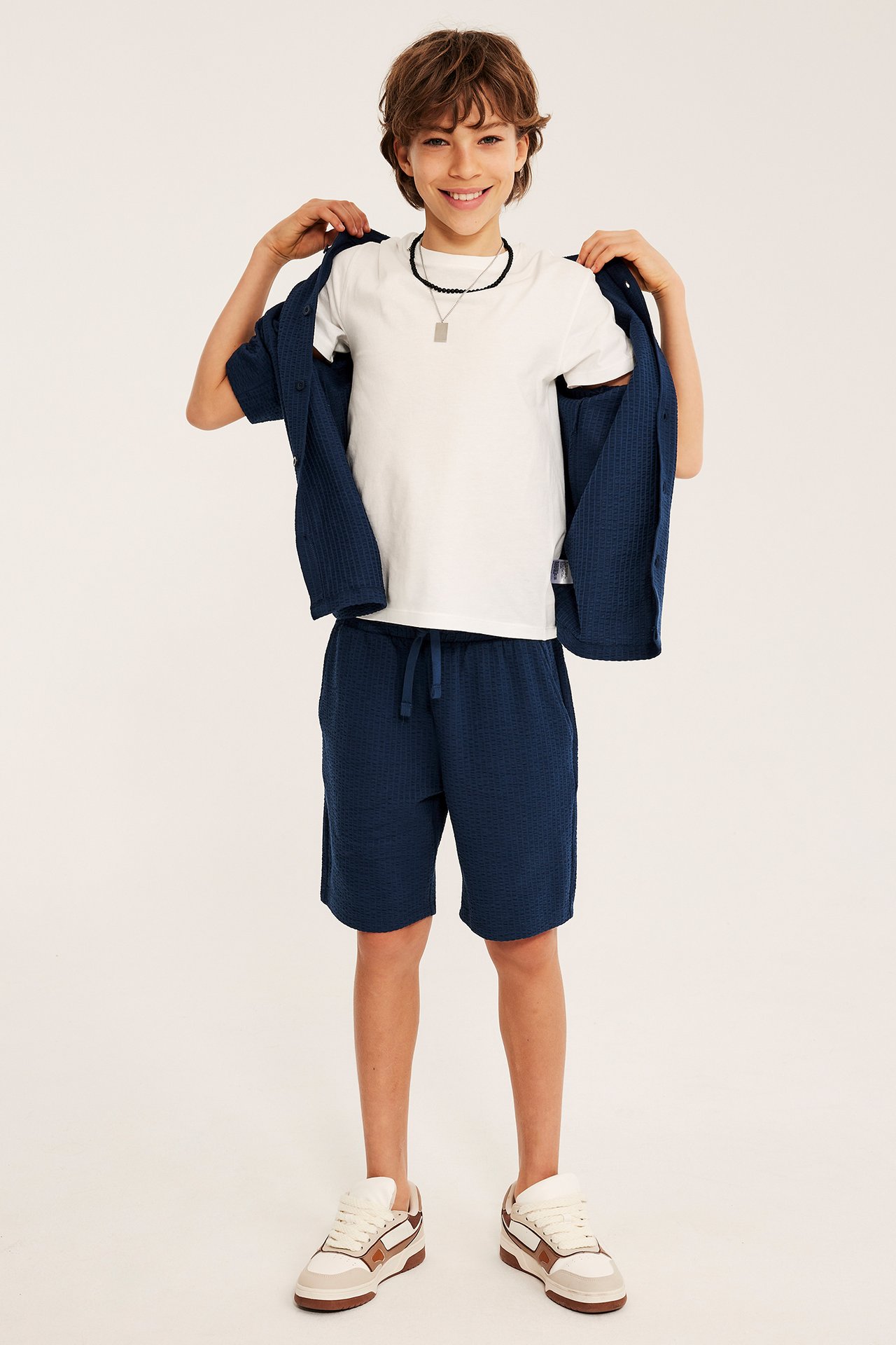 Widok z przodu: Dziecko w granatowych szortach z dżerseju seersucker, białym T-shircie, otwartej niebieskiej koszuli i trampkach.