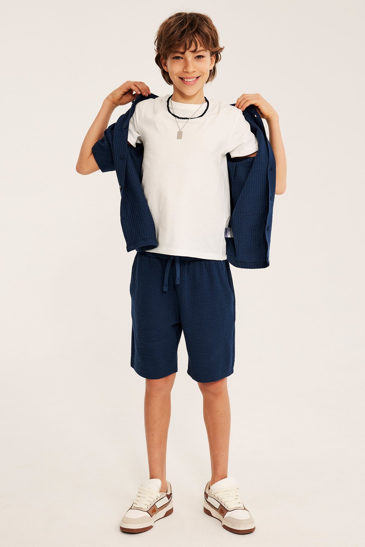 Widok z przodu: Dziecko w granatowych szortach z dżerseju seersucker, białym T-shircie, otwartej niebieskiej koszuli i trampkach.