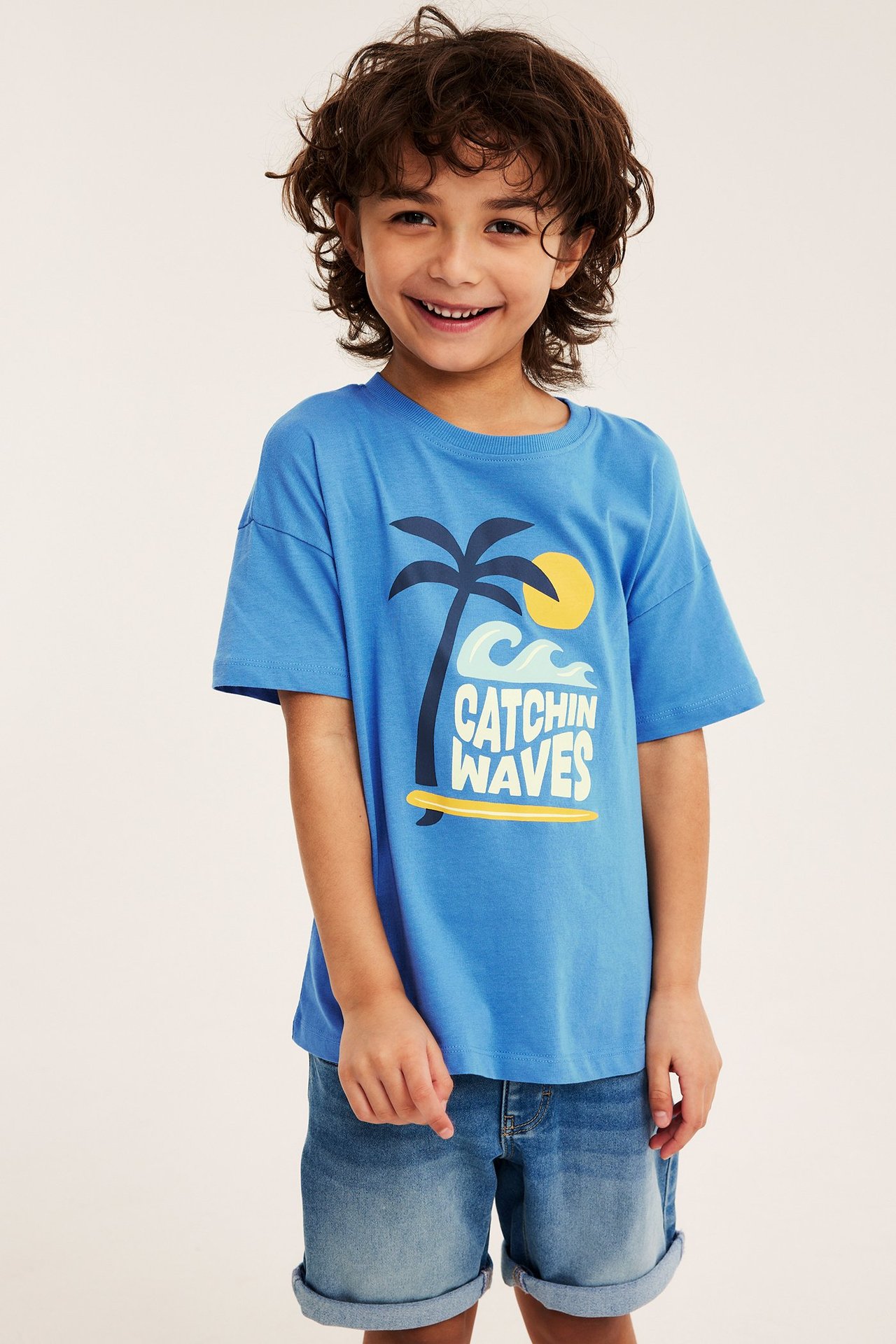 Edestäpäin hymyilevä lapsi sinisessä puuvillaisessa t-paidassa, jossa on 'Catchin Waves' -printti ja sinisissä farkkushortseissa.