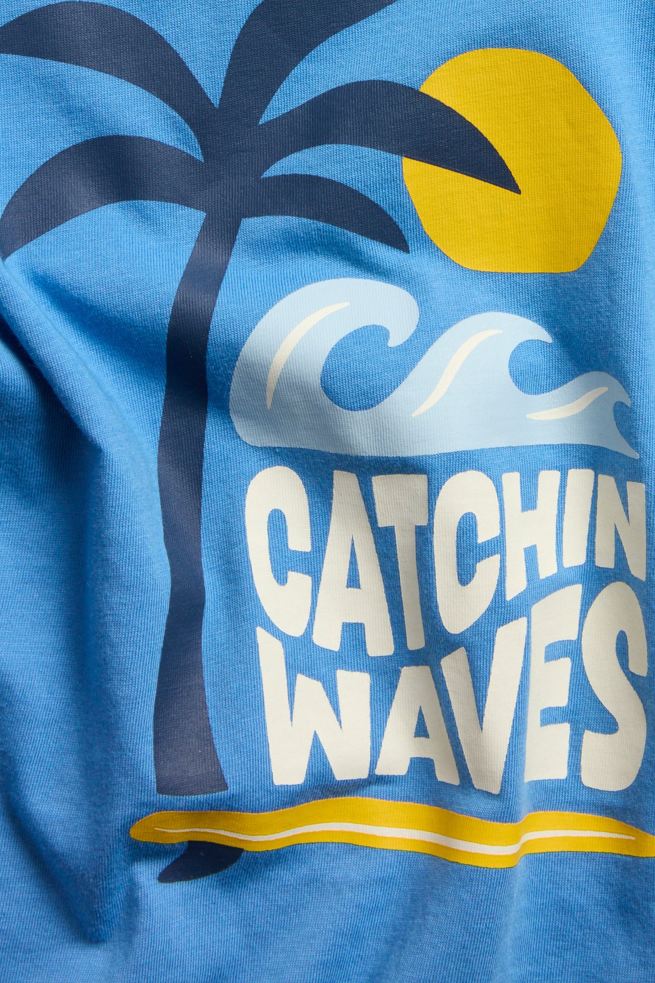 Zbliżenie z przodu: niebieska bawełniana koszulka dziecięca z nadrukiem palmy, słońca i „Catchin Waves”.
