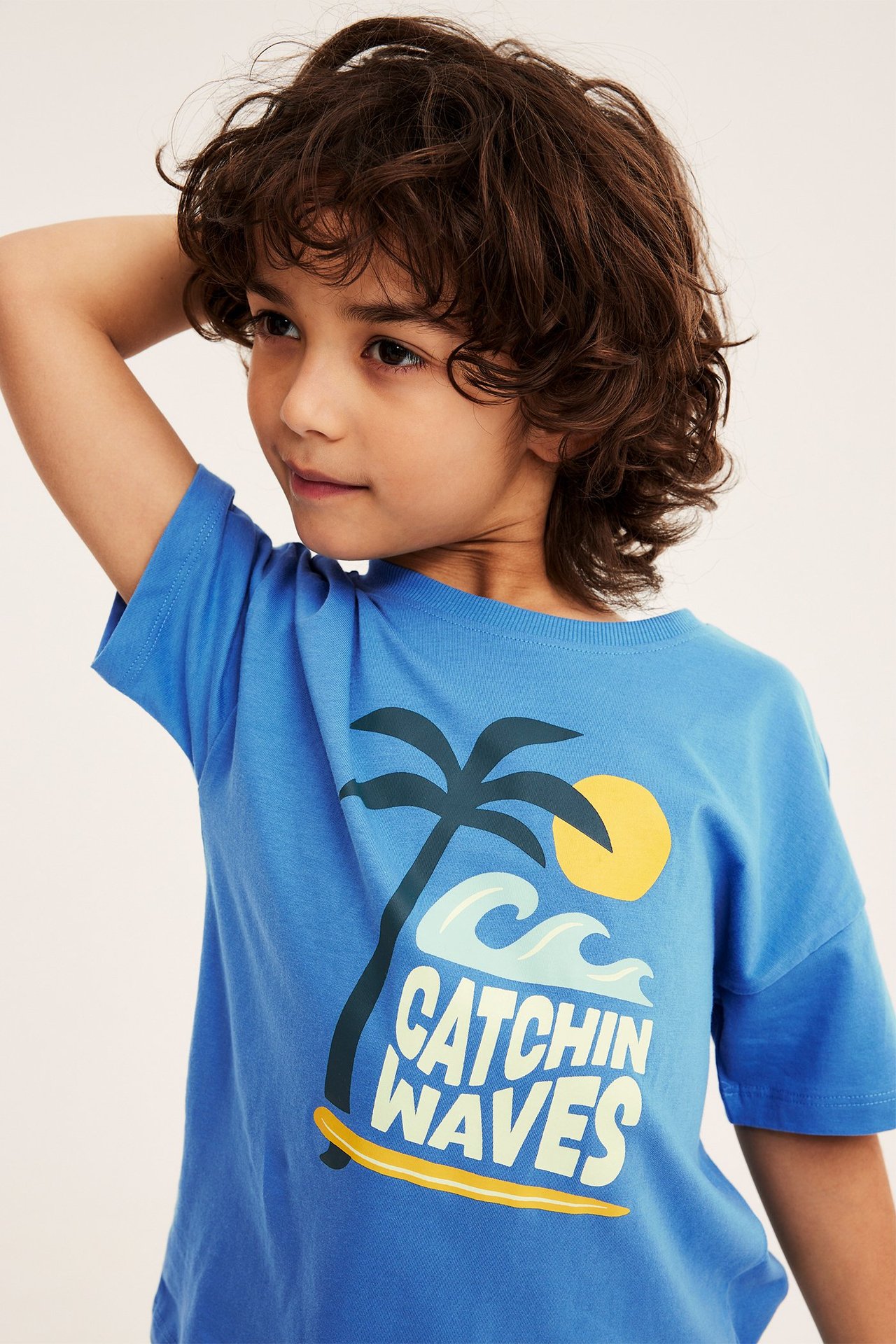 Widok z przodu, dziecko w niebieskiej bawełnianej koszulce z nadrukiem „Catchin' Waves” i ściągaczowym dekoltem.