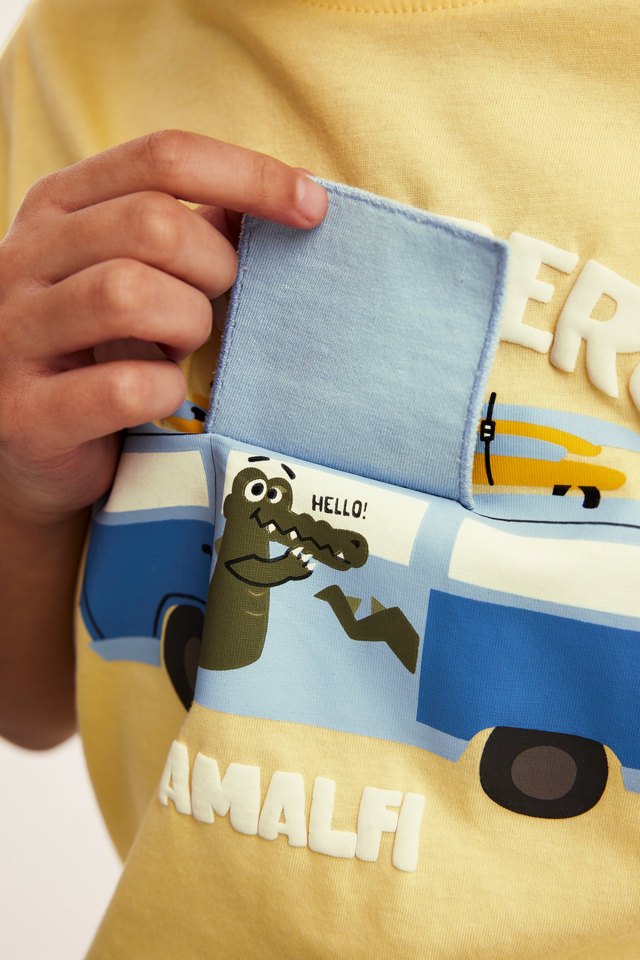 Närbild: Barns gul oversize t-shirt i bomull med krokodiltryck, en hand lyfter fliken.