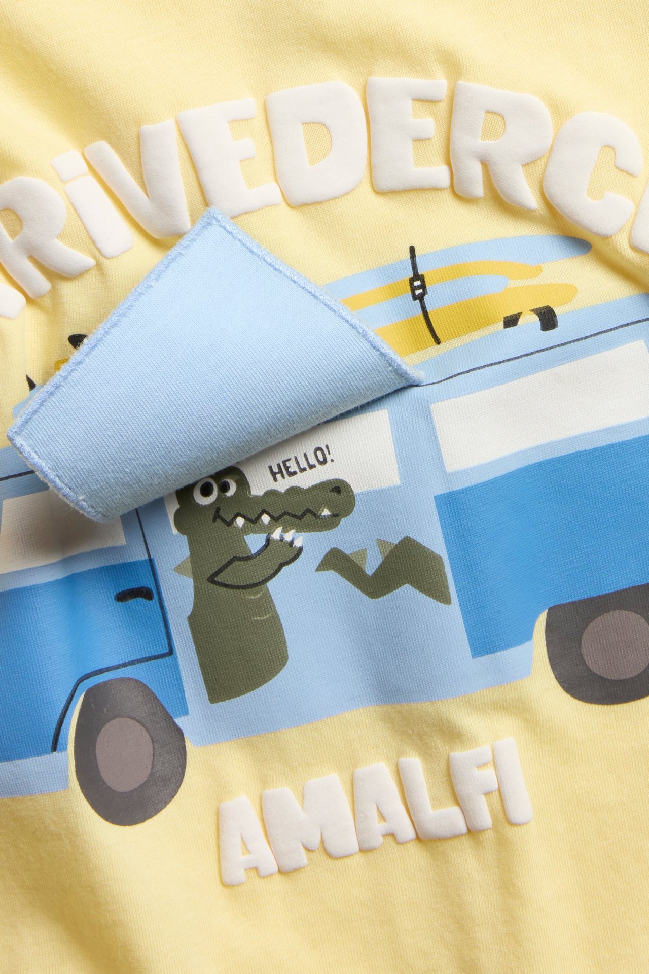 Zbliżenie: Żółty bawełniany t-shirt oversize z nadrukiem niebieskiego vana, krokodyla, wypukłymi literami i interaktywną klapką.